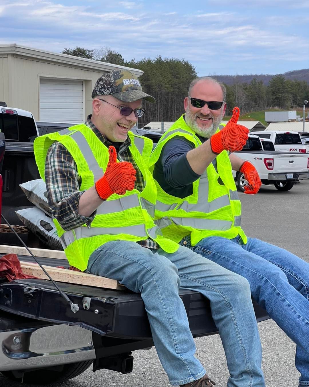 Two workers in neon vests and orange gloves sit on a truck bed, smiling and giving thumbs up.