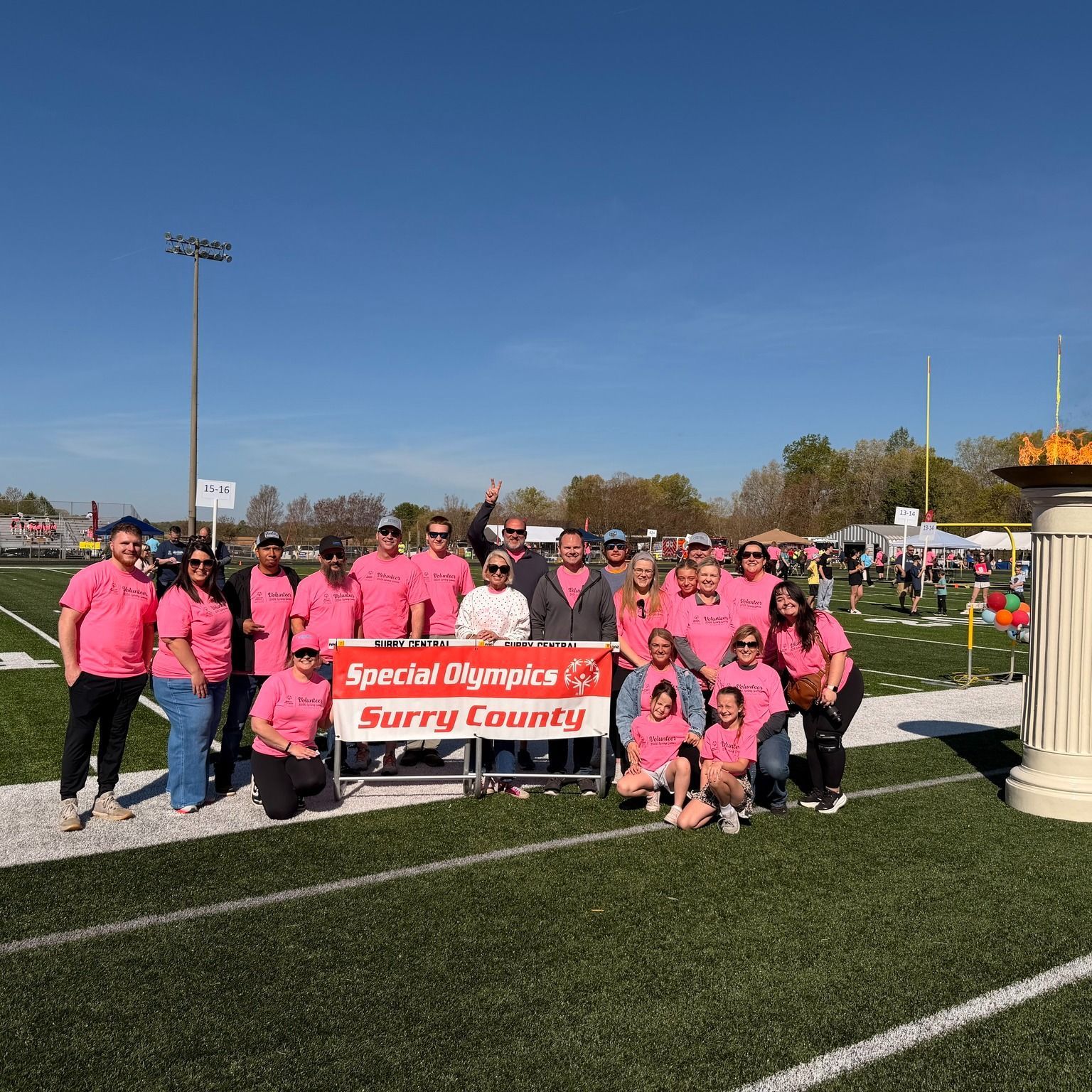 Group of employees in pink shirts posing with a banner at a sports field on a sunny day.