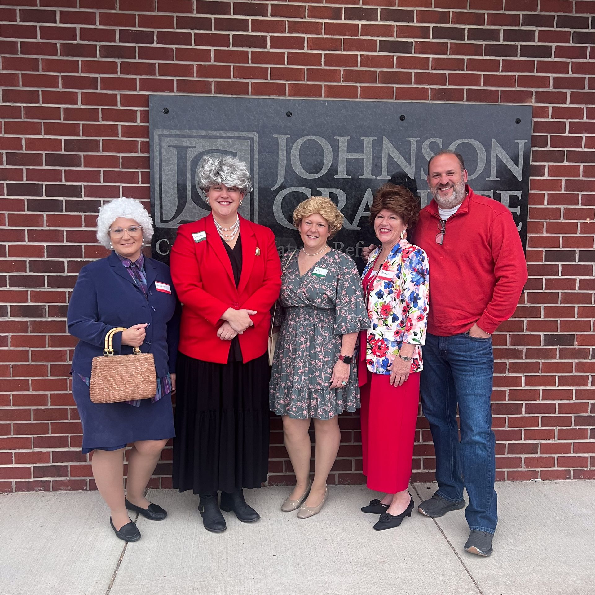 Five people posing in front of a brick wall and sign, two wearing red jackets and one holding a purse.