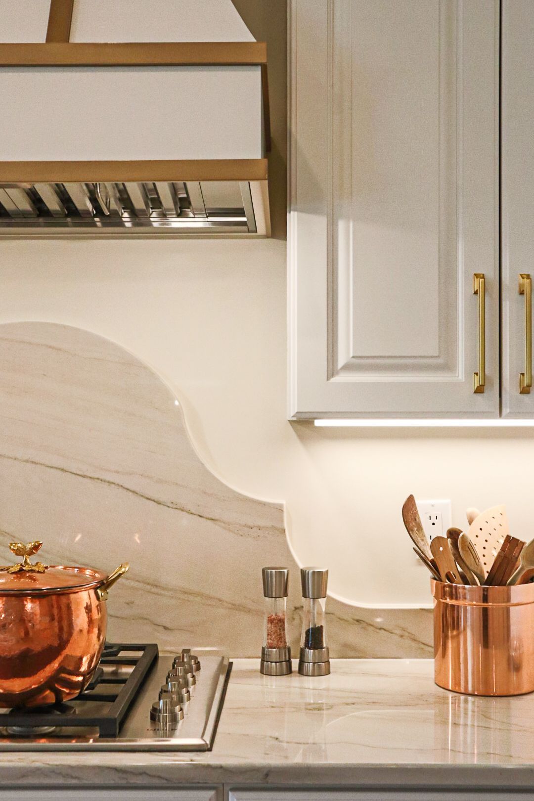 Modern kitchen with white cabinets, copper cookware, utensils, and a marble countertop.