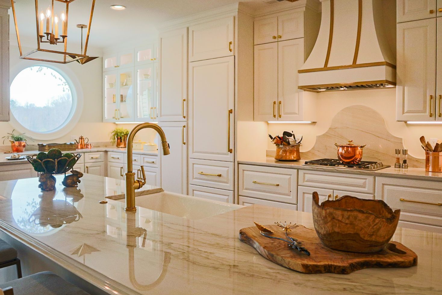 Bright modern kitchen with marble island, gold faucet, white cabinets, and round window.