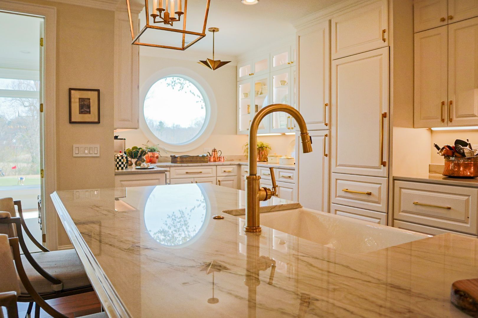 Bright modern kitchen with marble island, gold faucet, white cabinets, and round window.