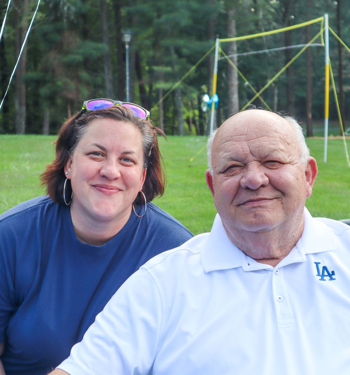 Two people smiling outdoors, wearing blue and white shirts, with a sports field in the background