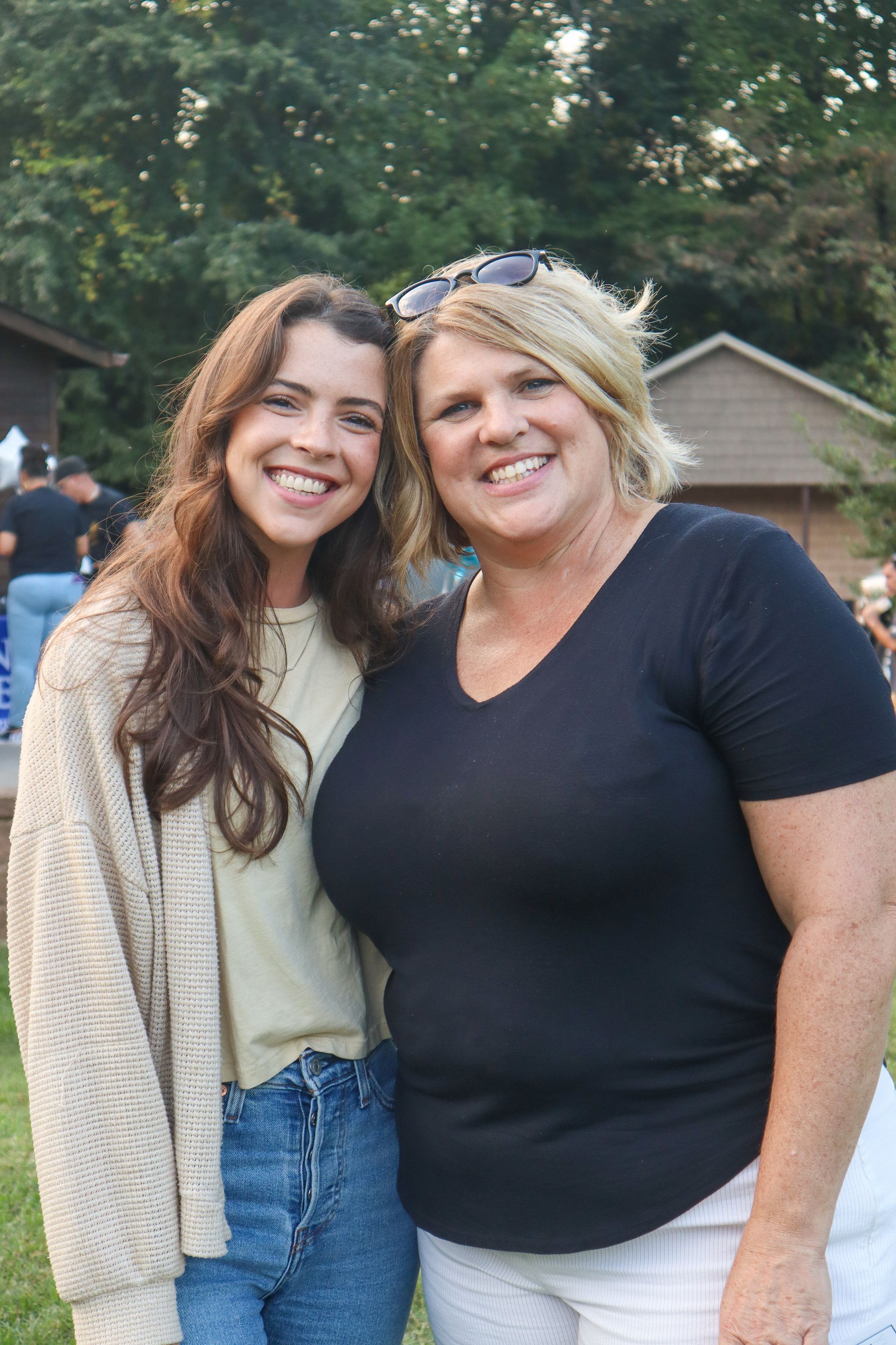 Two women smiling outdoors, standing close together with trees and houses in the background
