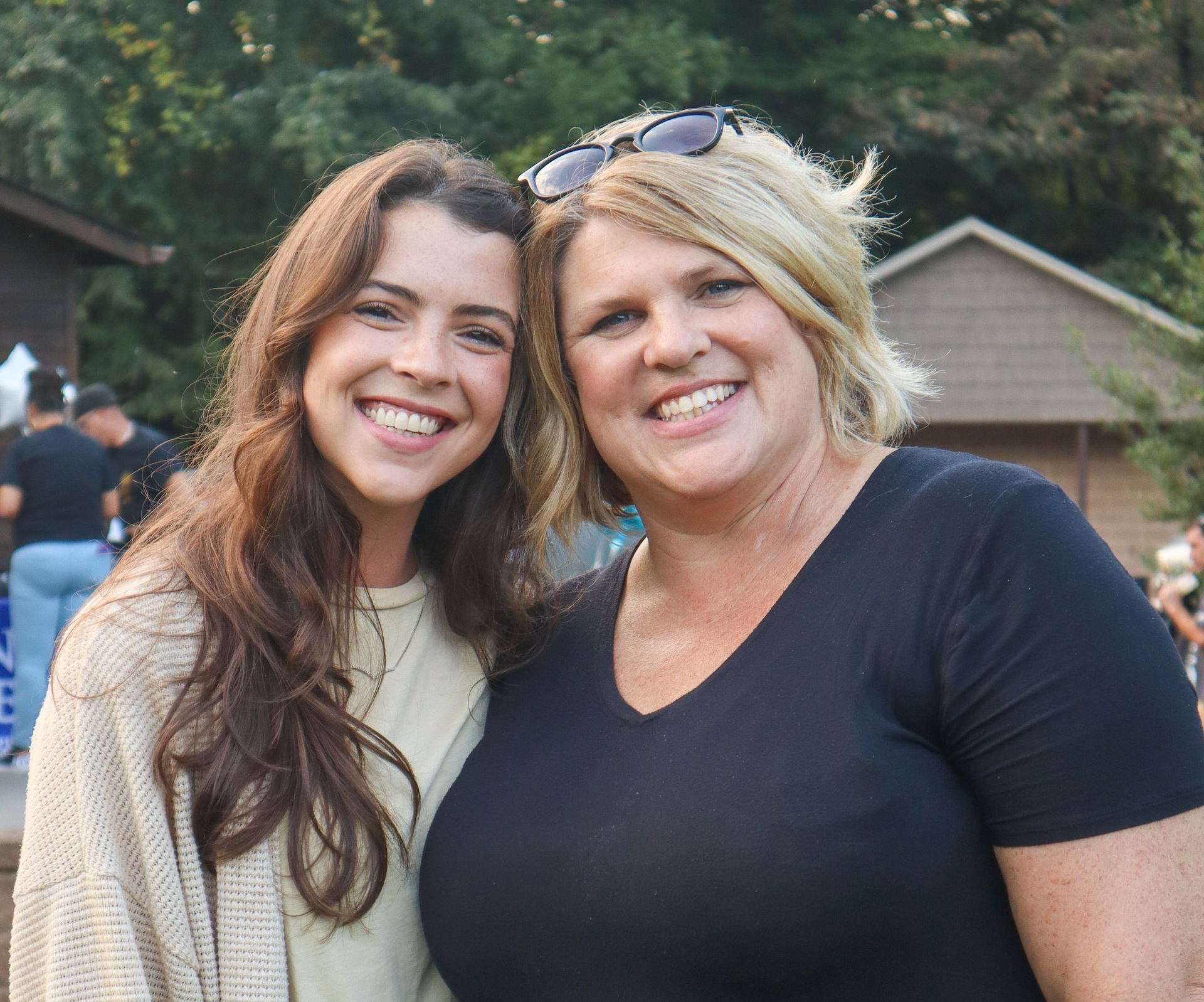 Two smiling women standing outdoors, one with long brown hair and one blonde in a black shirt.