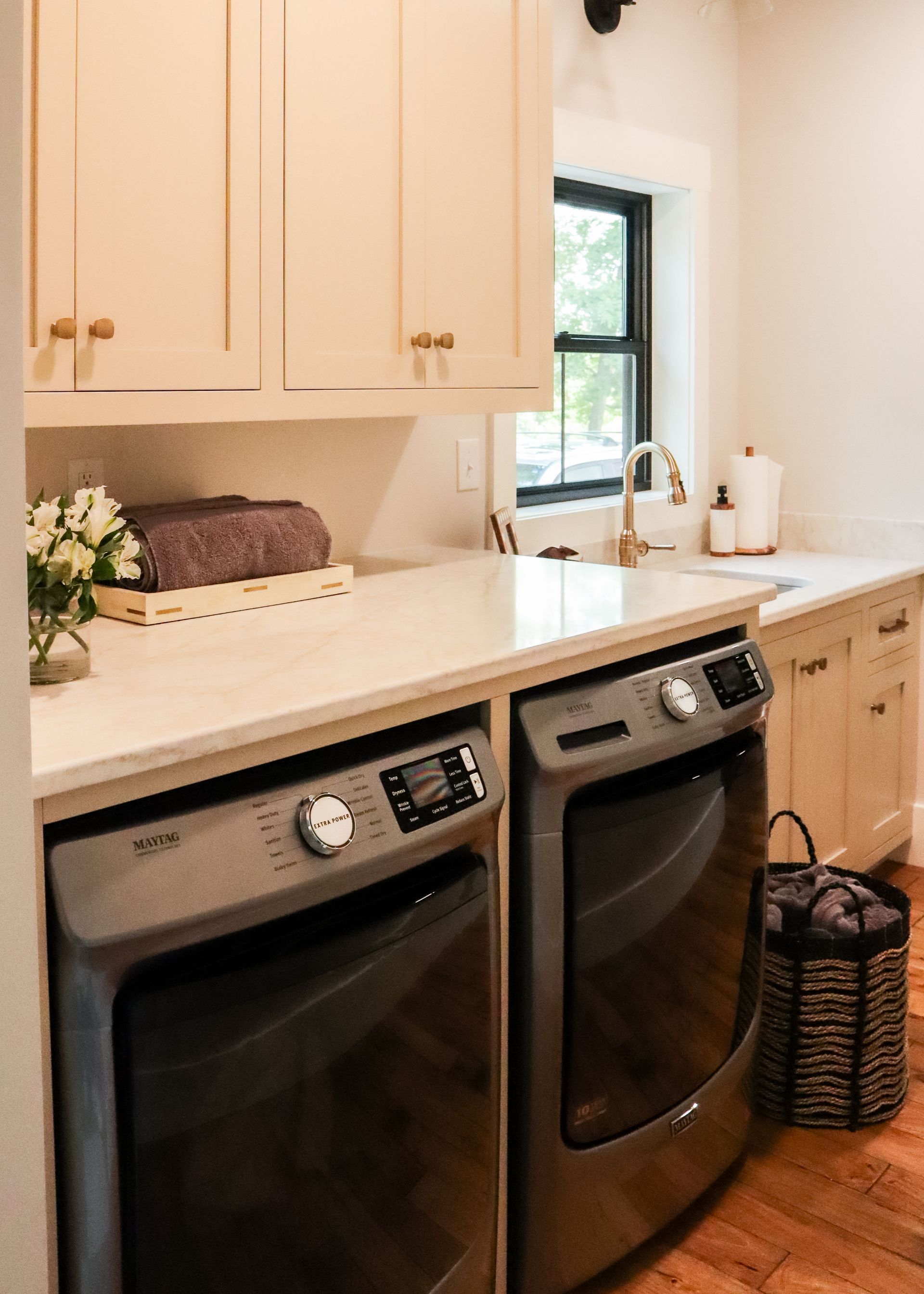 Modern laundry room with two front-loading washer-dryer units, white cabinets, and a window over the countertop