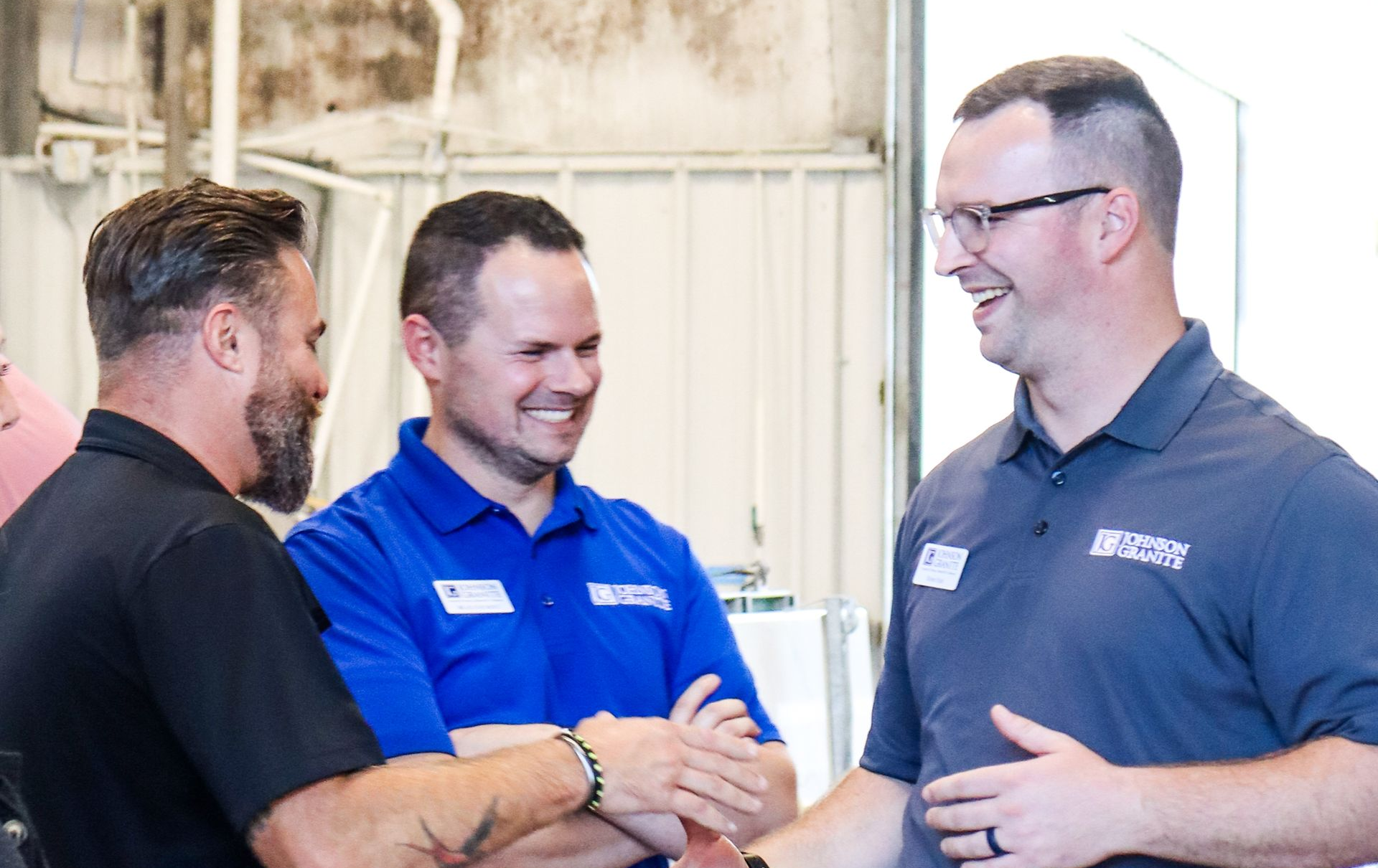 Three men in blue shirts smiling and shaking hands indoors at an event.