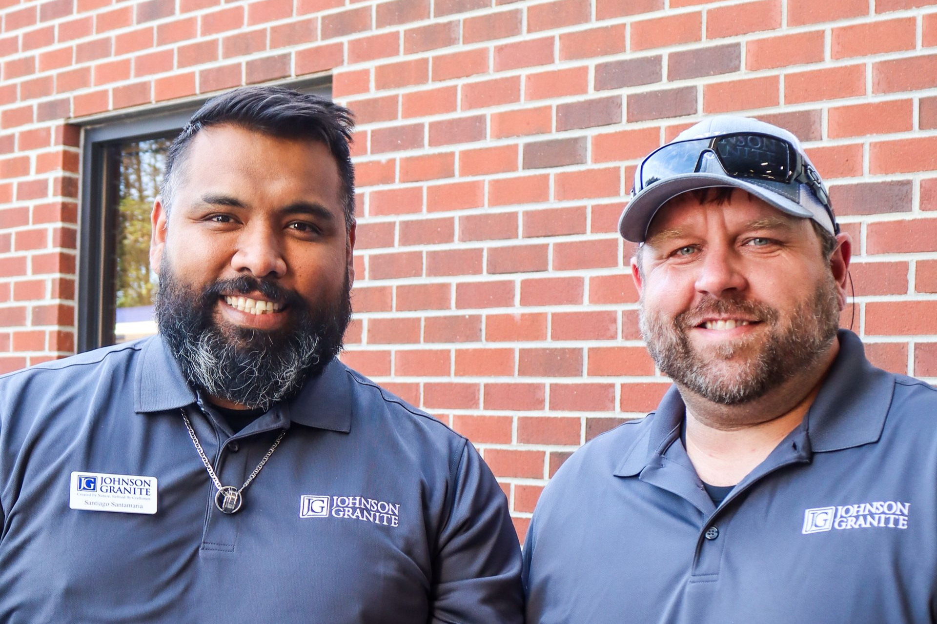 Two smiling employees in navy shirts standing in front of a brick wall