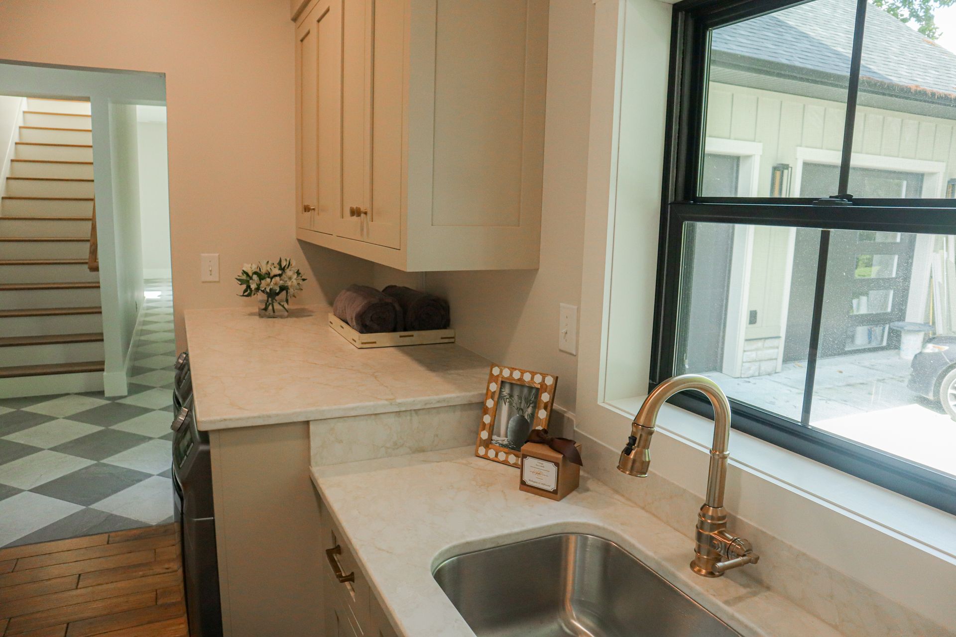 Bright kitchen sink and counter beside large window, with beige cabinets and a doorway to stairs.