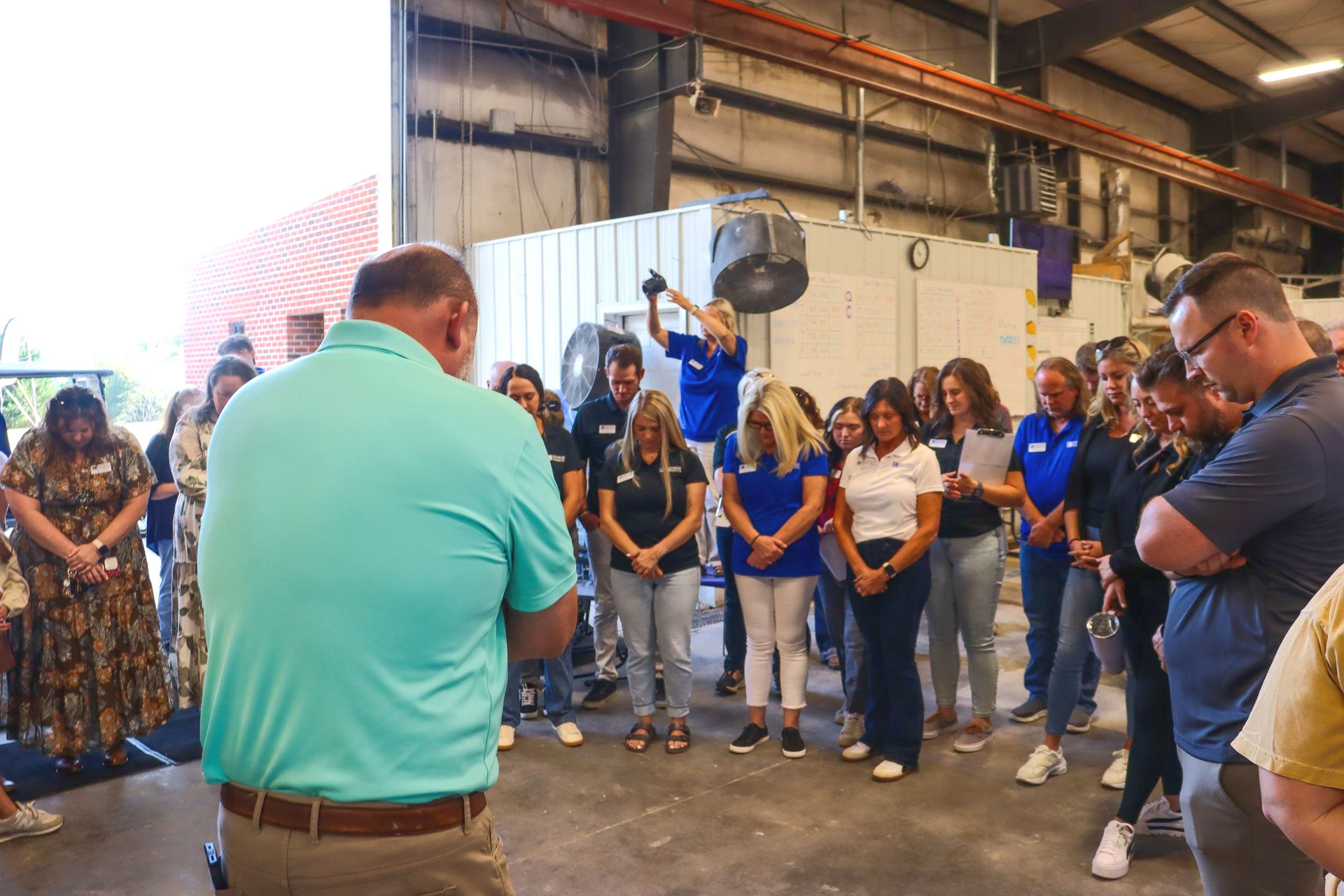 People standing in a semicircle indoors, heads bowed in a quiet gathering with one man facing them.