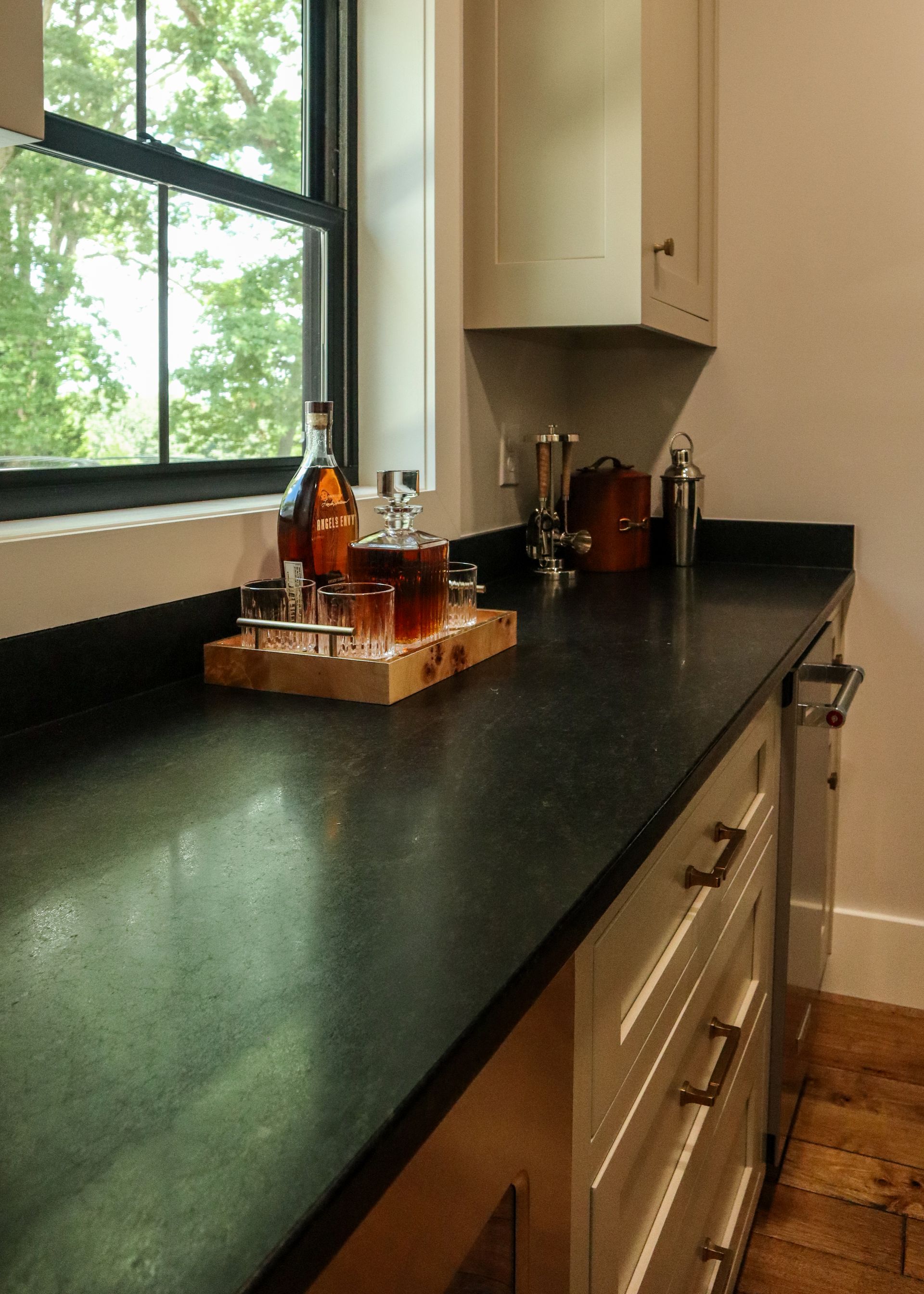 Kitchen countertop by a window with bottles and a tray, dark granite surface, wood floor and cabinets