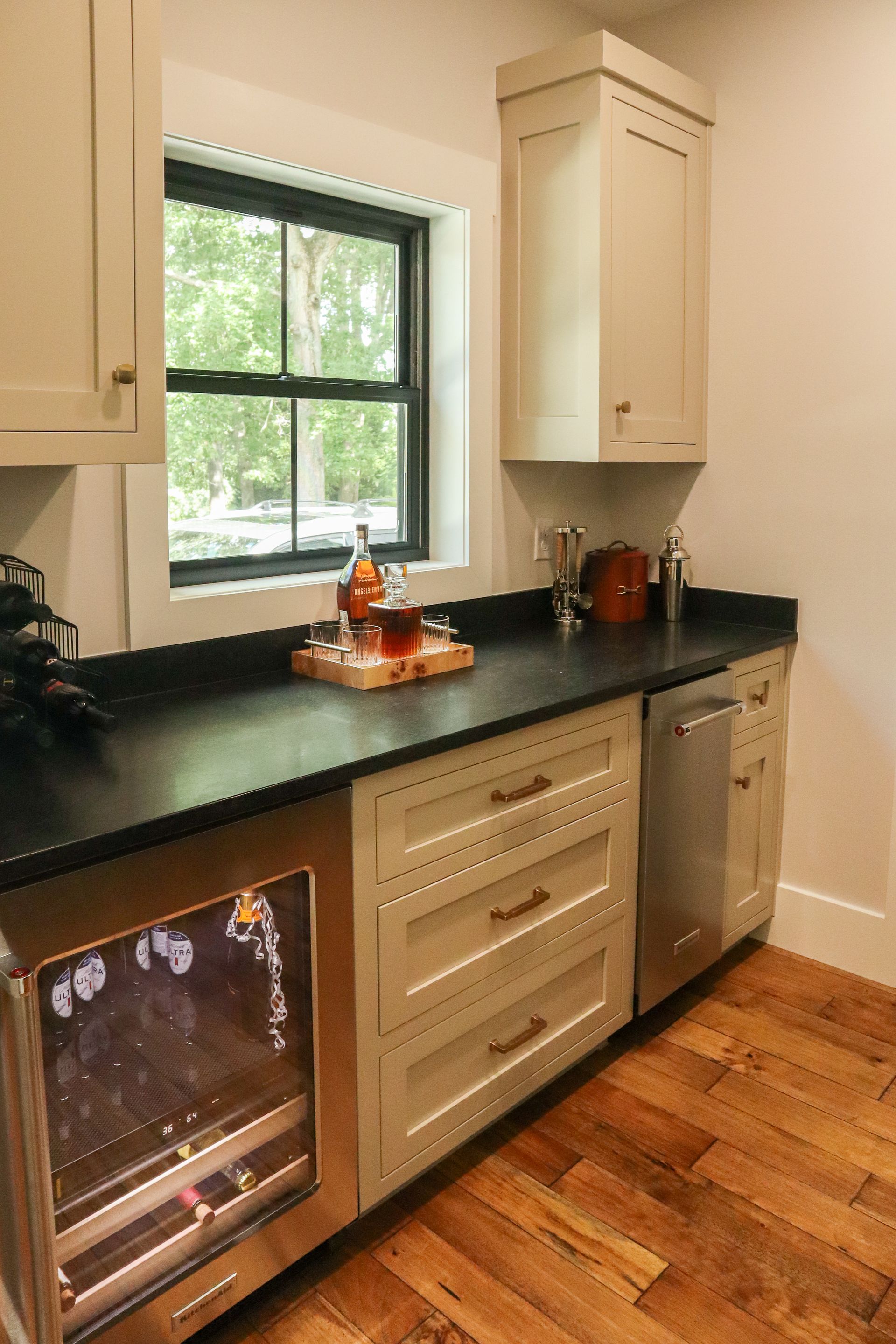 Kitchen counter with dark countertop, window, cabinets, and stainless steel dishwasher above wood floor.