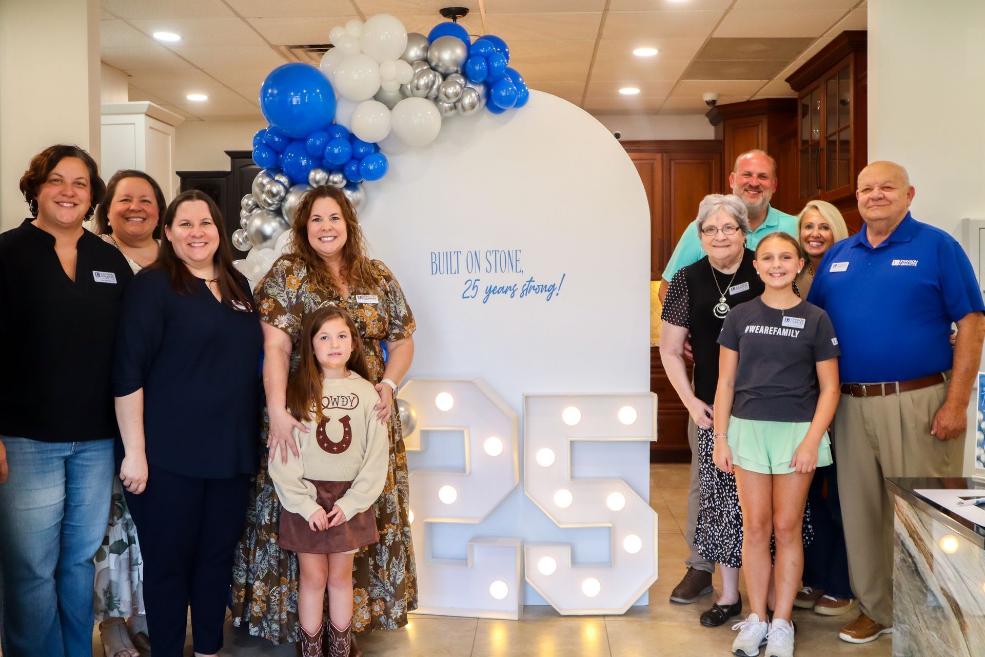Johnson family posing at a blue-and-white balloon backdrop with a large lit “25” display indoors celebrating 25 years in business