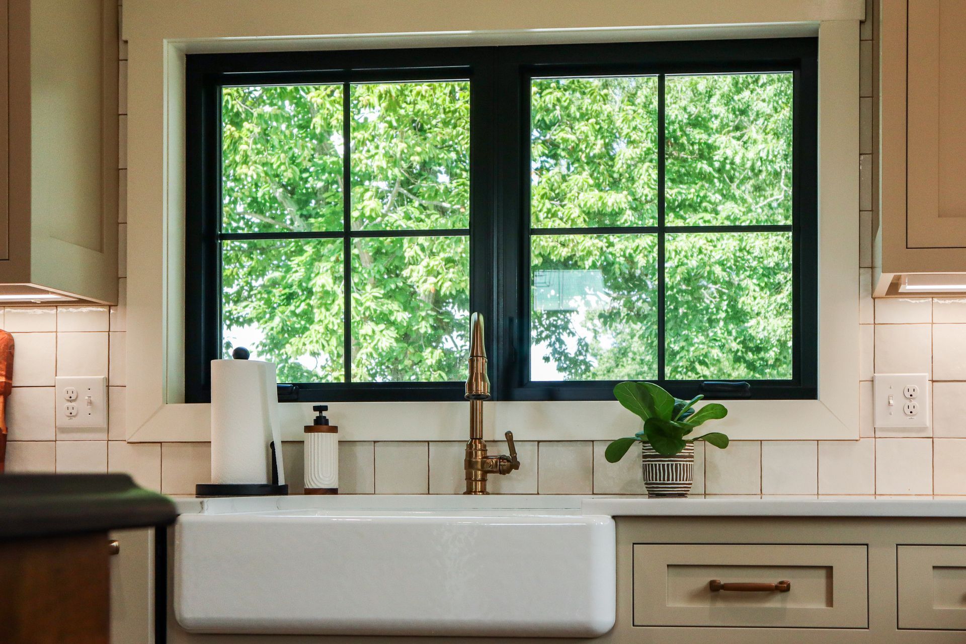 Kitchen sink beneath large window with black frames, white countertop, and potted plant, overlooking green trees