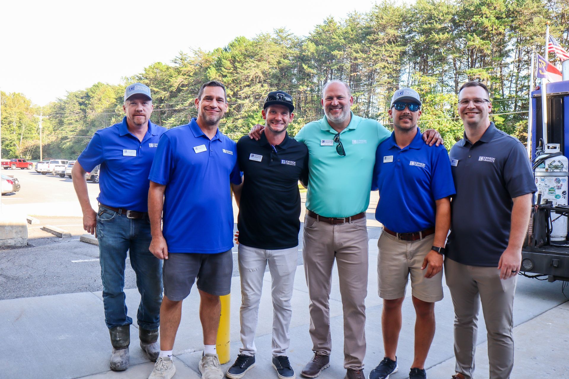 Six JGI employees standing outdoors in front of trees, smiling and posing together.