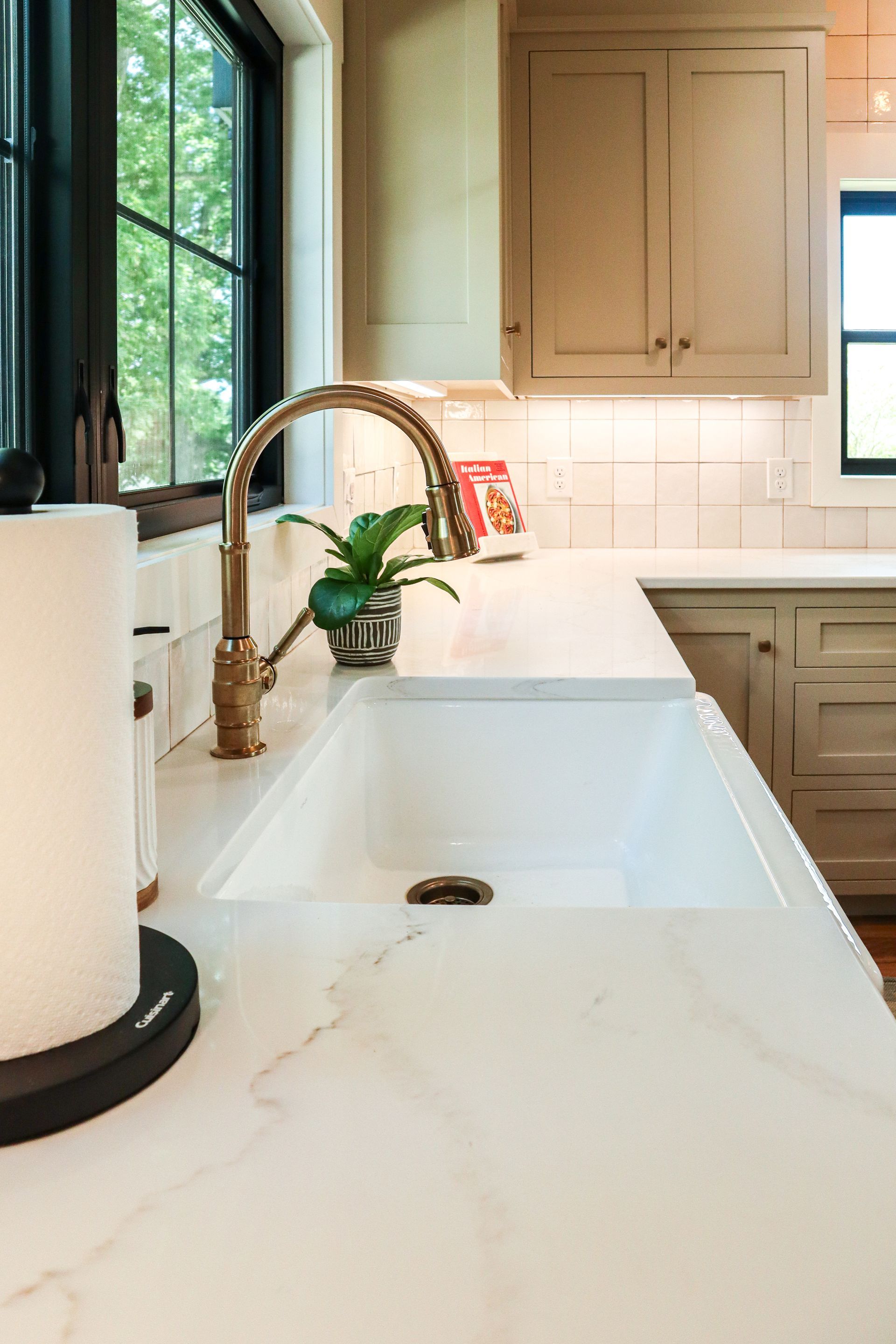Bright white kitchen sink with gold faucet by a window, surrounded by beige cabinets and countertops.