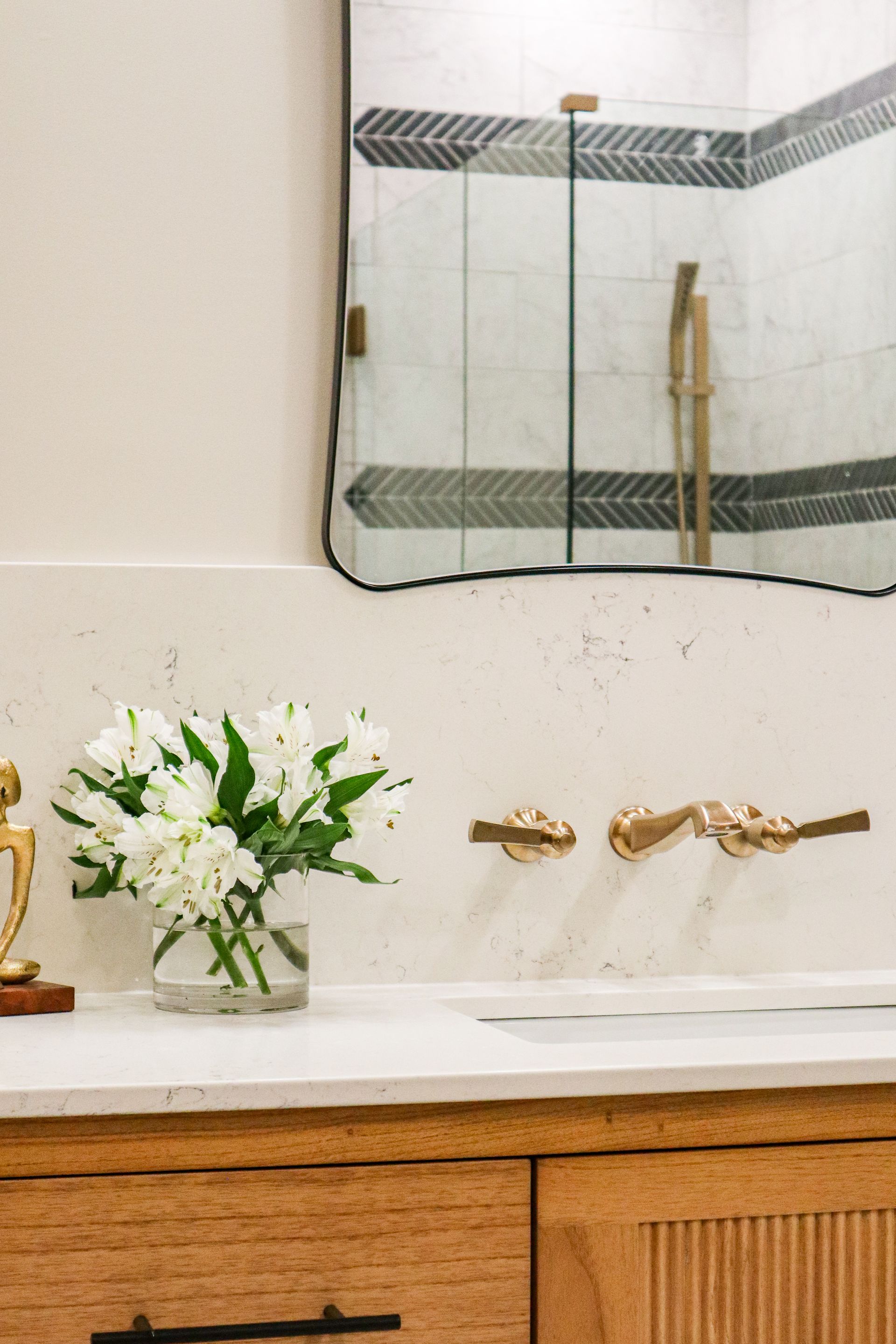 Bathroom vanity with gold faucet, white sink, white flowers, and mirror reflecting a shower.
