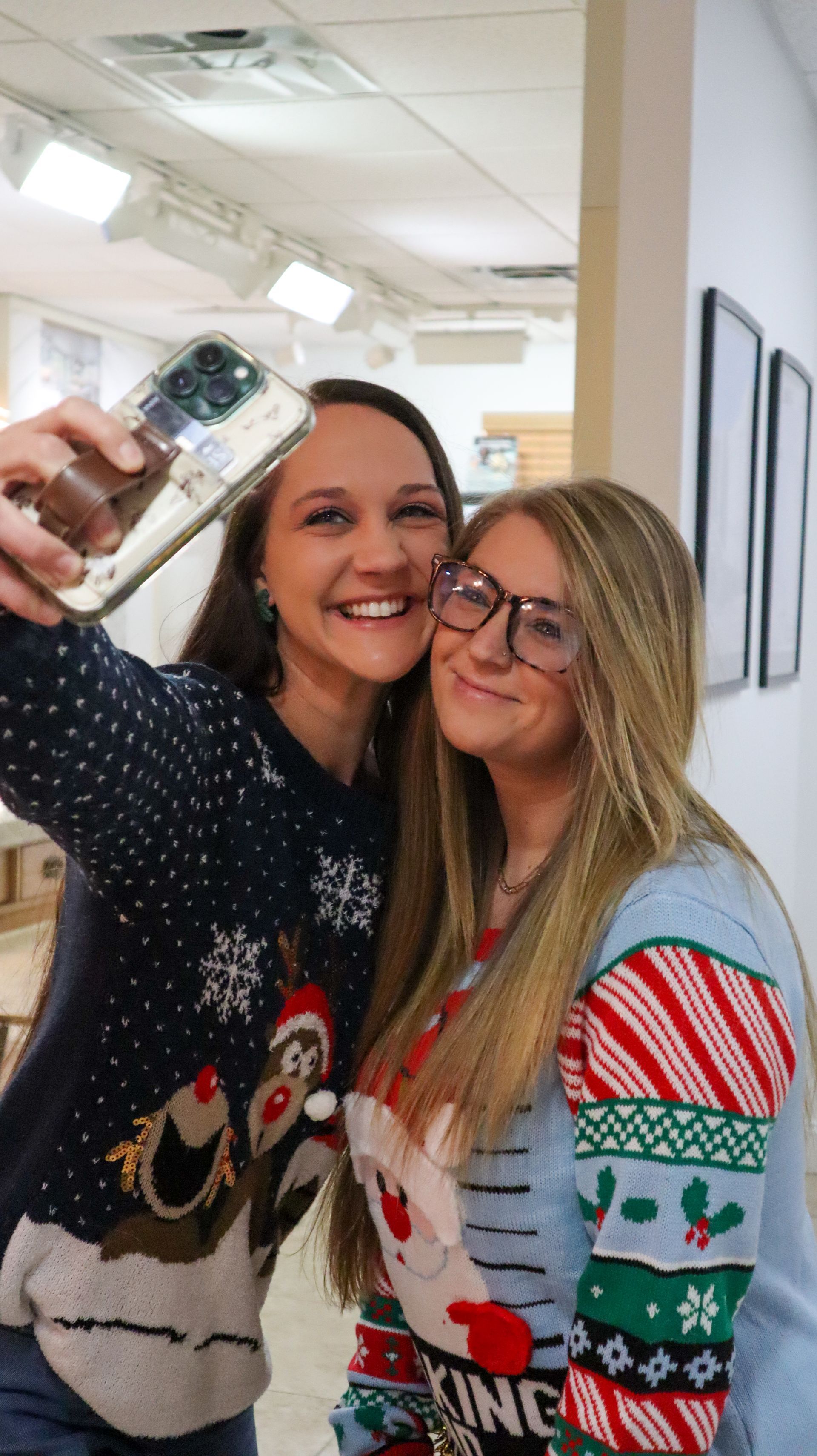 Two smiling women taking a selfie indoors, one holding a phone, both in festive sweaters.