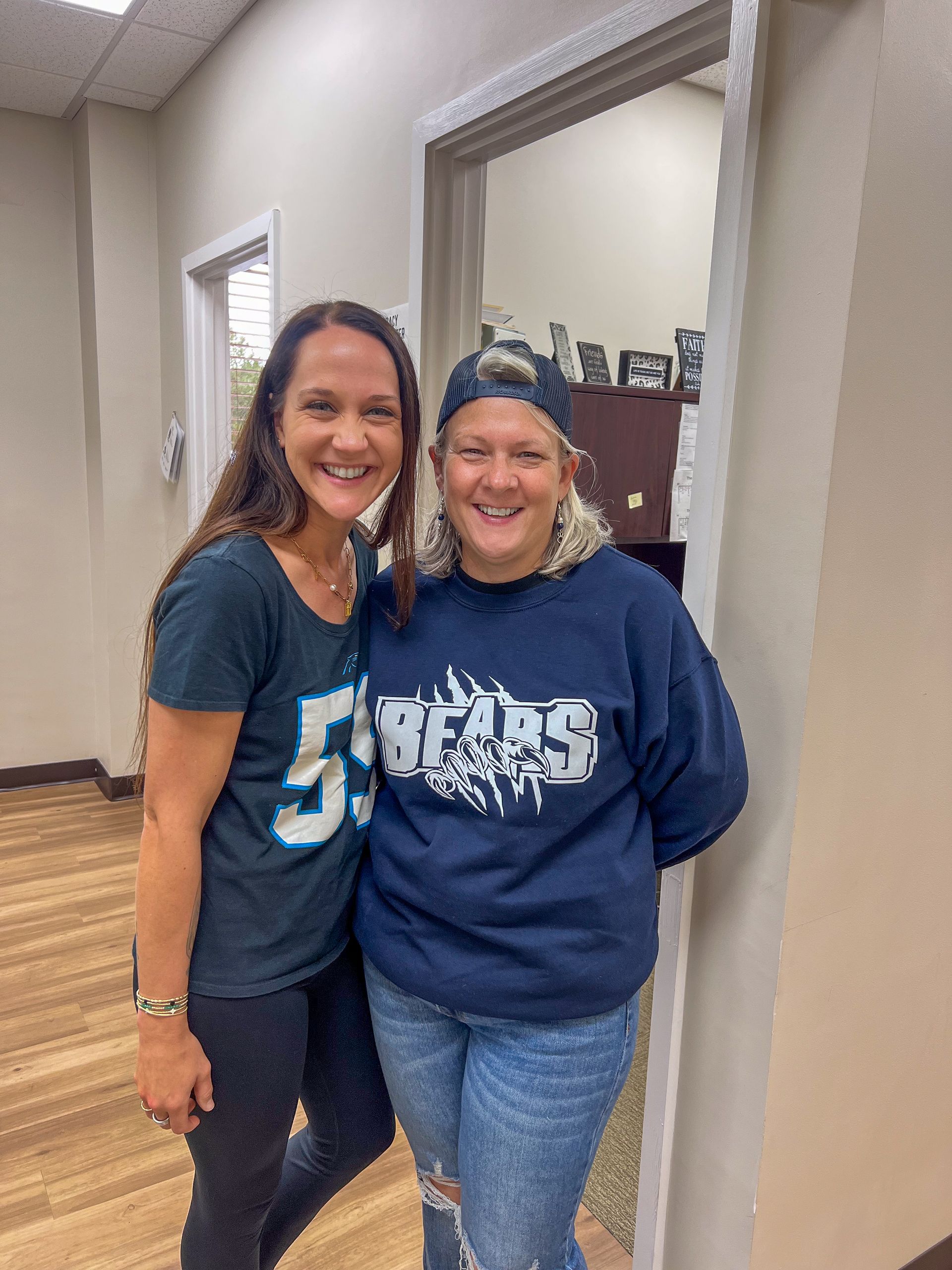 Two smiling women standing together indoors in a hallway, wearing navy shirts and jeans.