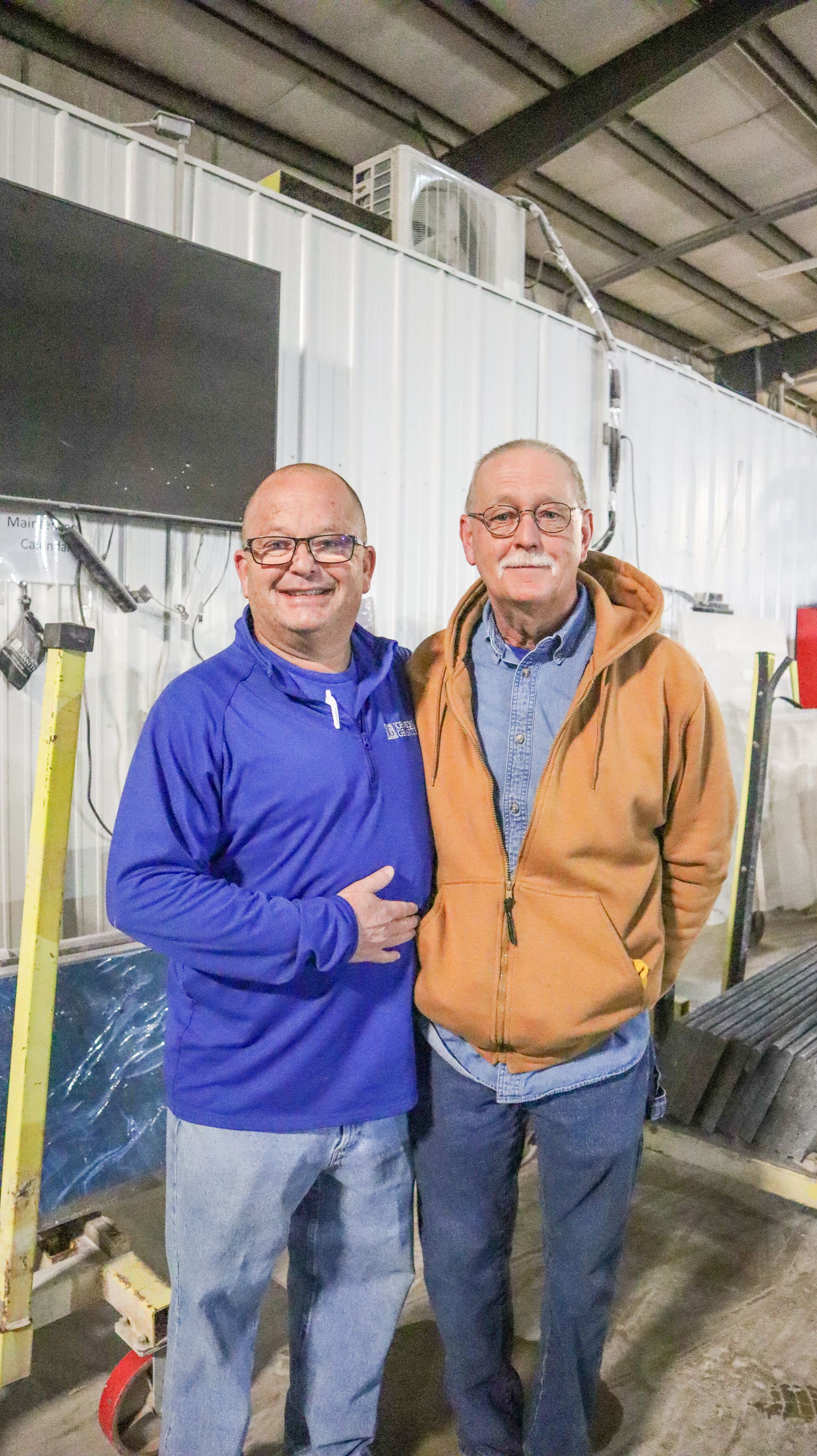 Two smiling men standing together in a workshop, one in a blue pullover and one in an orange jacket.