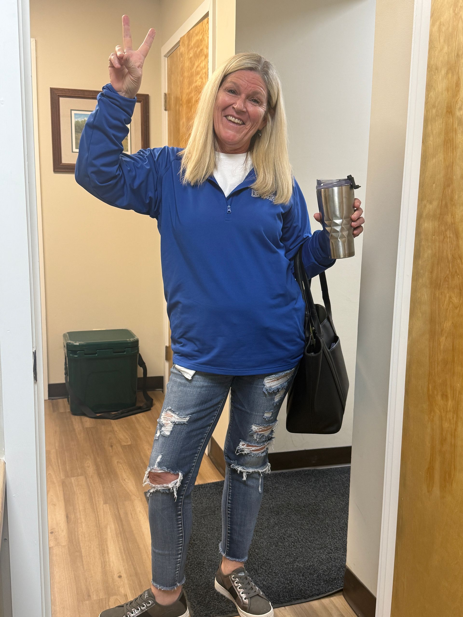 Smiling person in a blue sweater and ripped jeans waves while holding a tumbler in a hallway