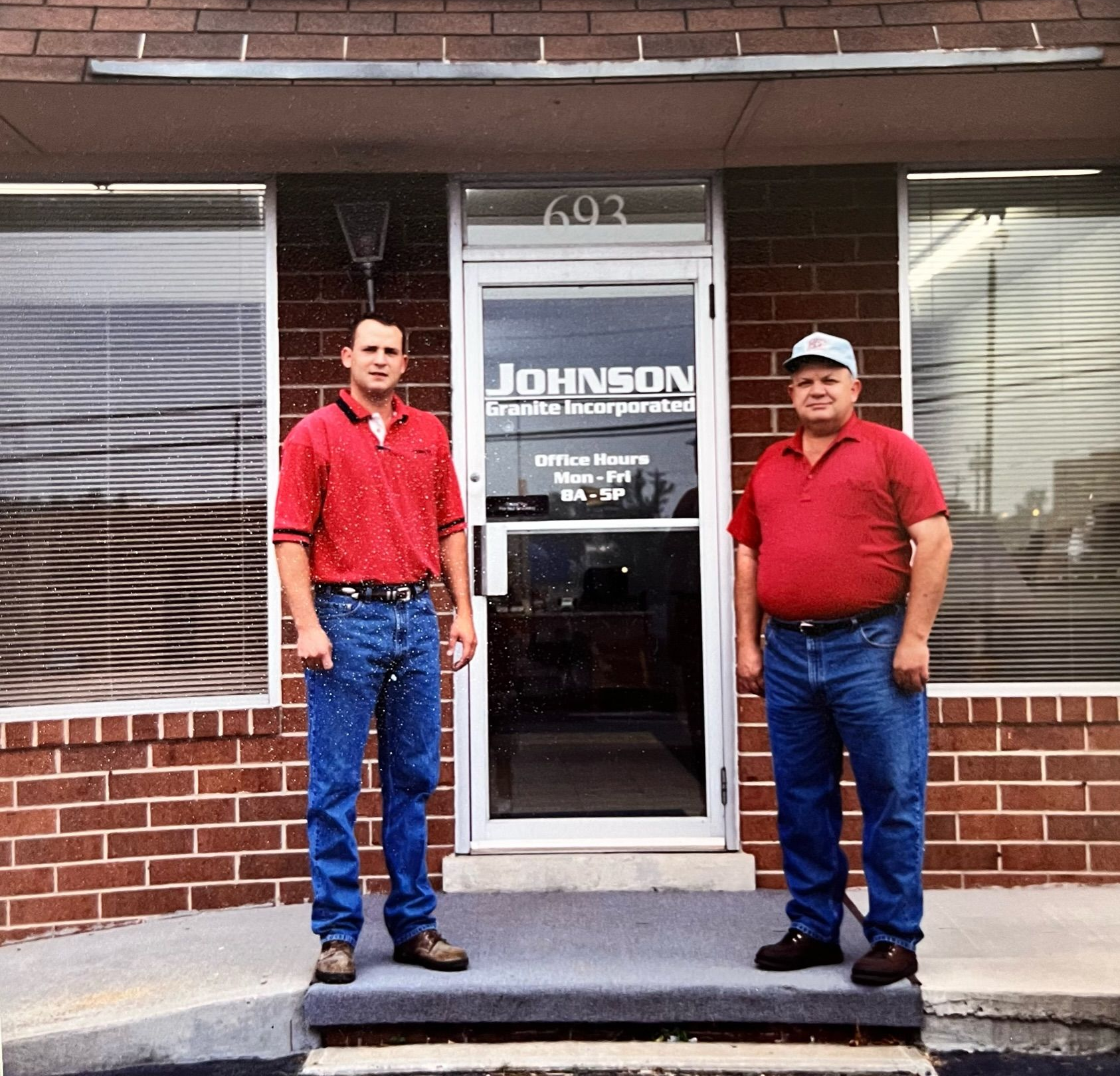 Larry & Brian Johnson in red shirts stand outside Johnson office storefront with a glass door and brick facade.