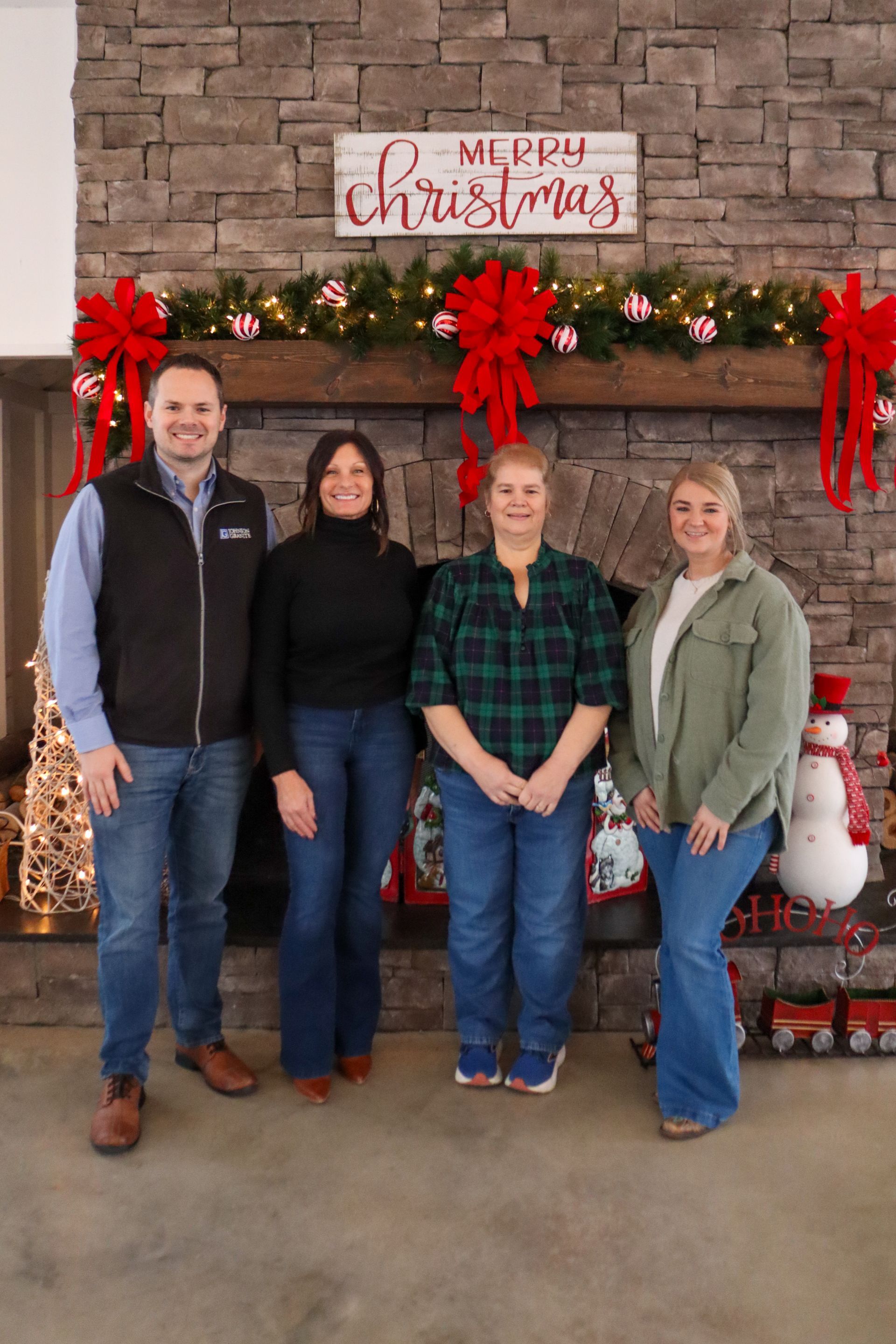 Four people posing in front of a holiday fireplace decorated with greenery and red bows.