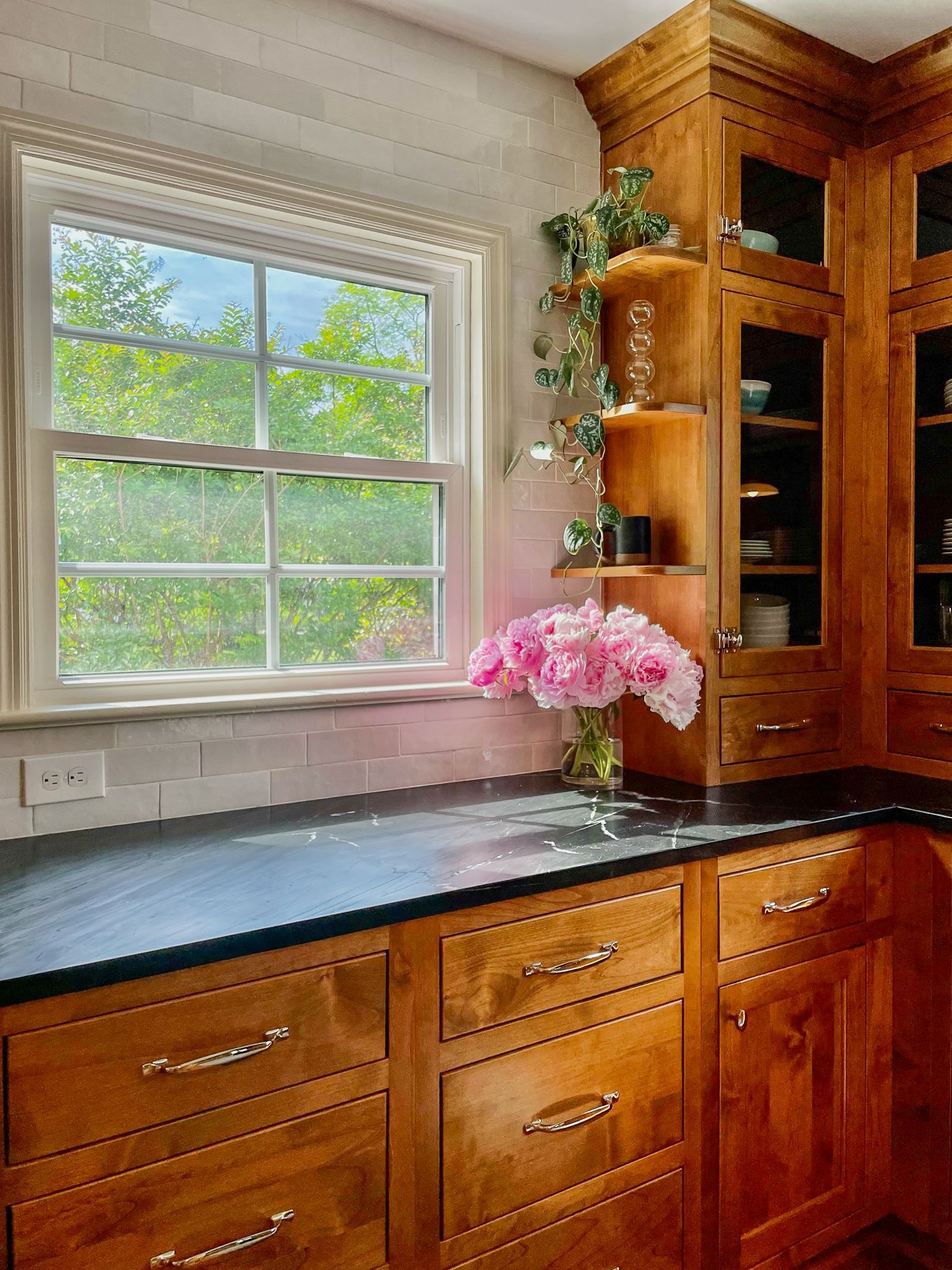 Kitchen with window, black countertop, wooden cabinets, and pink flowers by the sink.