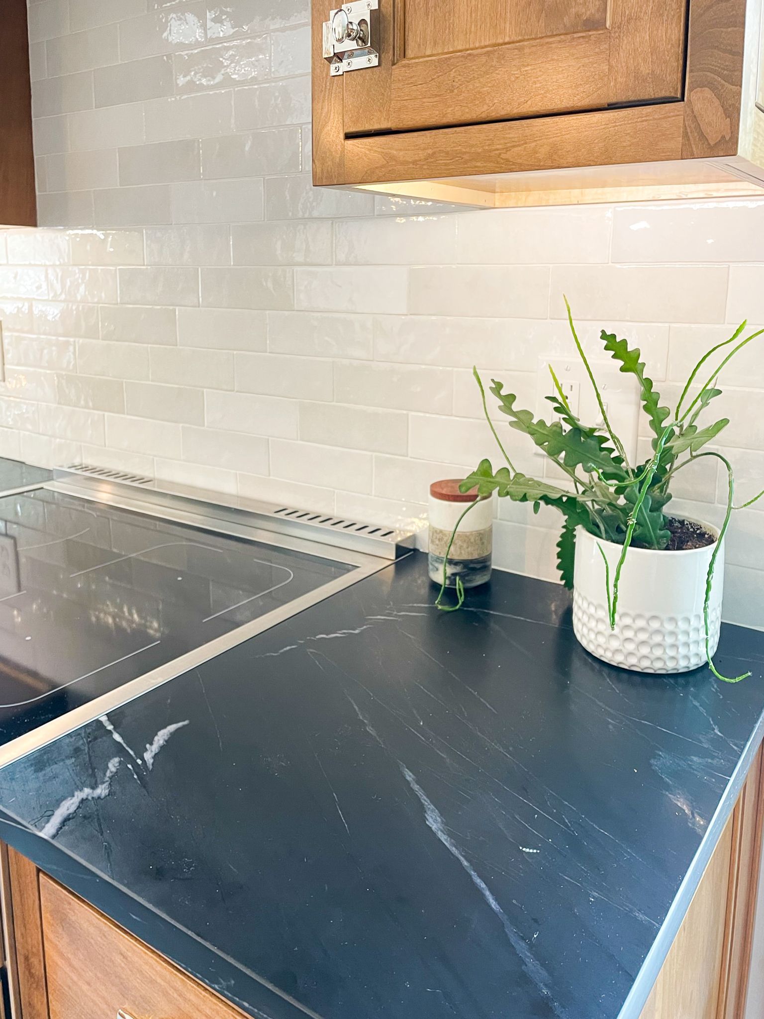 Modern kitchen counter with dark stone top, white tile backsplash, potted plant, and small jar near the stove top