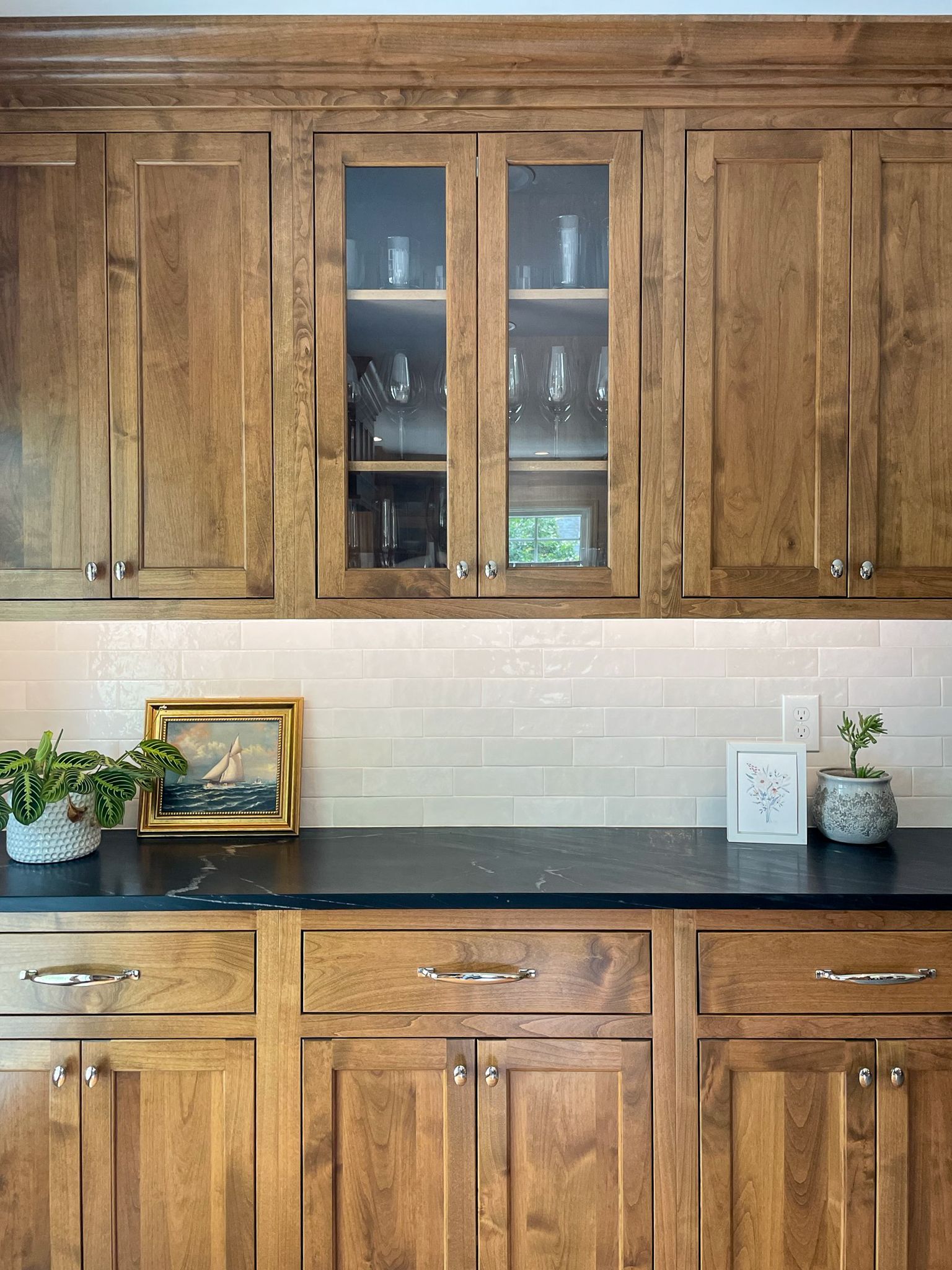 Wooden kitchen cabinets with glass-front center, black countertop, white tile backsplash, and small plants and framed art.