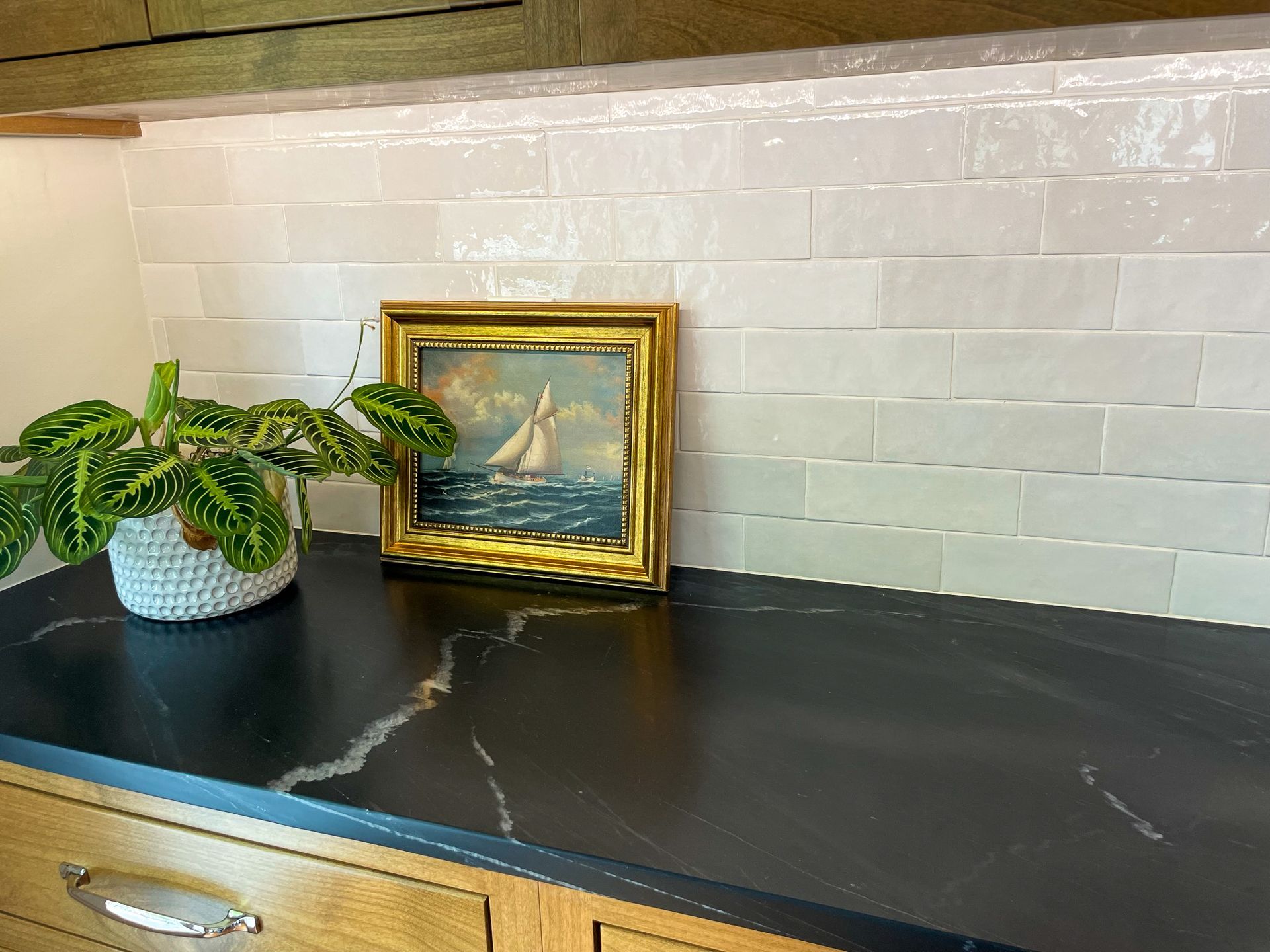 Small potted plant and framed sailboat painting on a black countertop against white tile backsplash