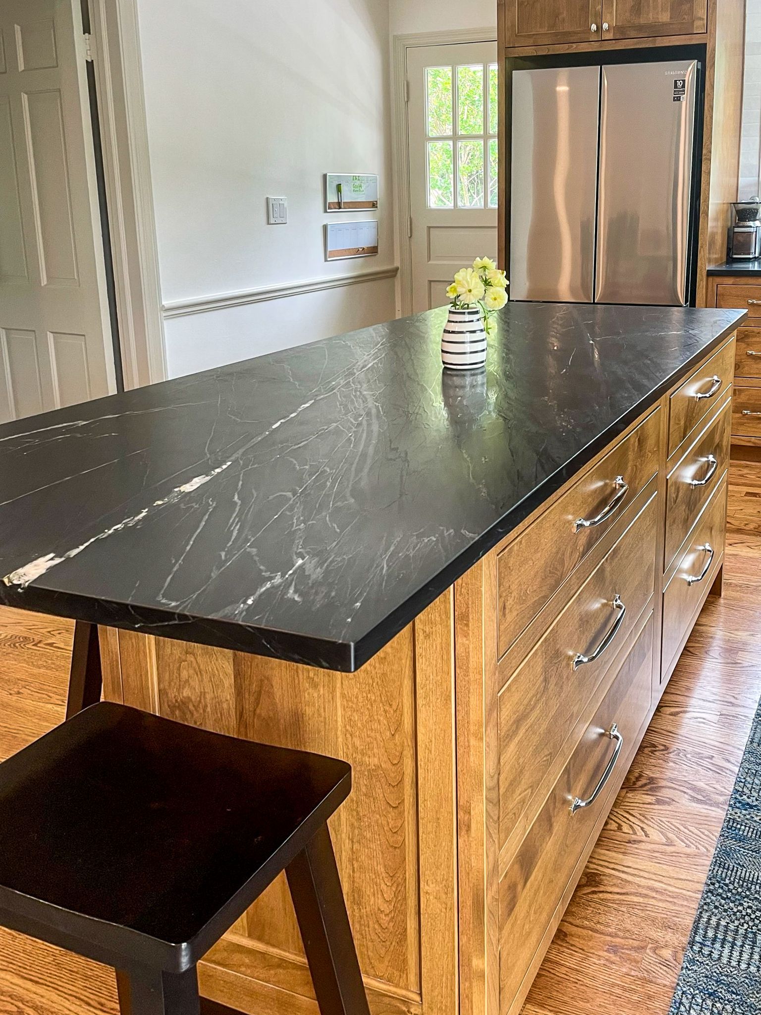 Kitchen island with black countertop, wood cabinets, a stool, and a stainless steel refrigerator in the background