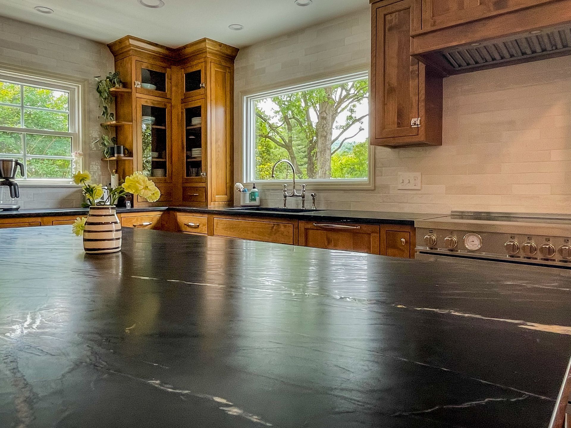 Modern kitchen with dark countertop, wooden cabinets, stainless stove, and large windows overlooking greenery