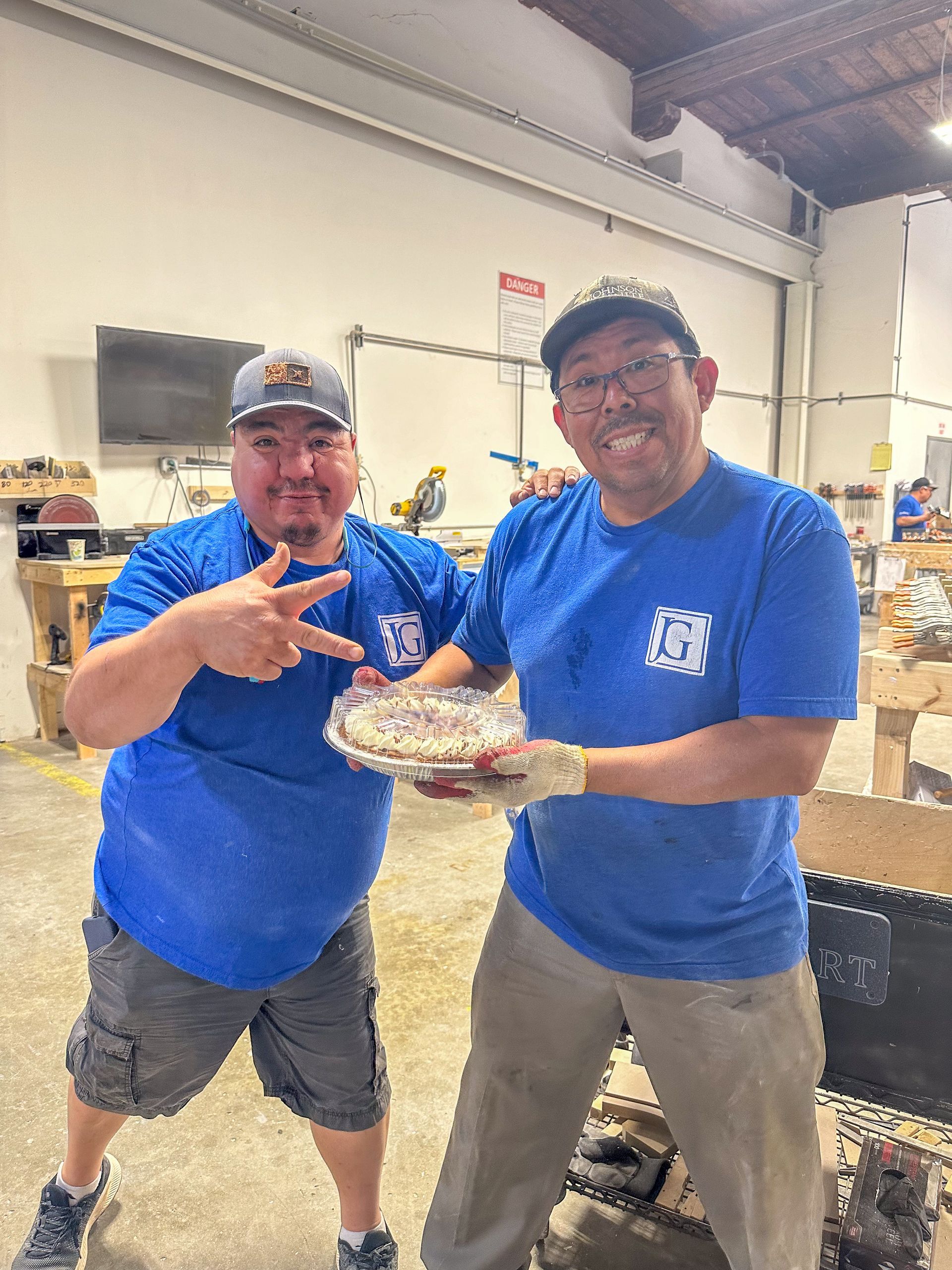 Two men in blue shirts hold a decorated cake in a workshop, smiling at the camera.
