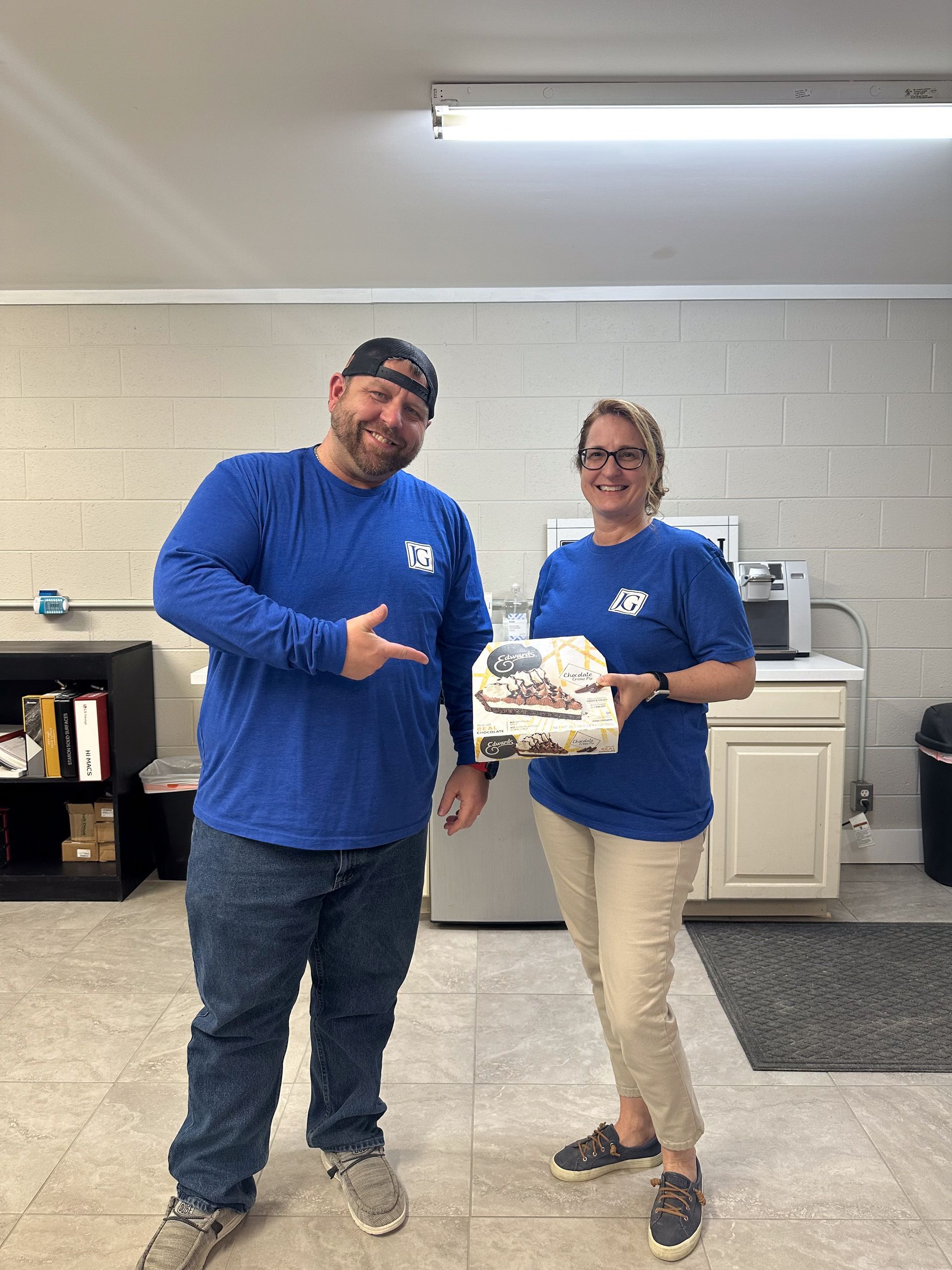 Two people in blue shirts smiling and holding a wrapped cake in a break room
