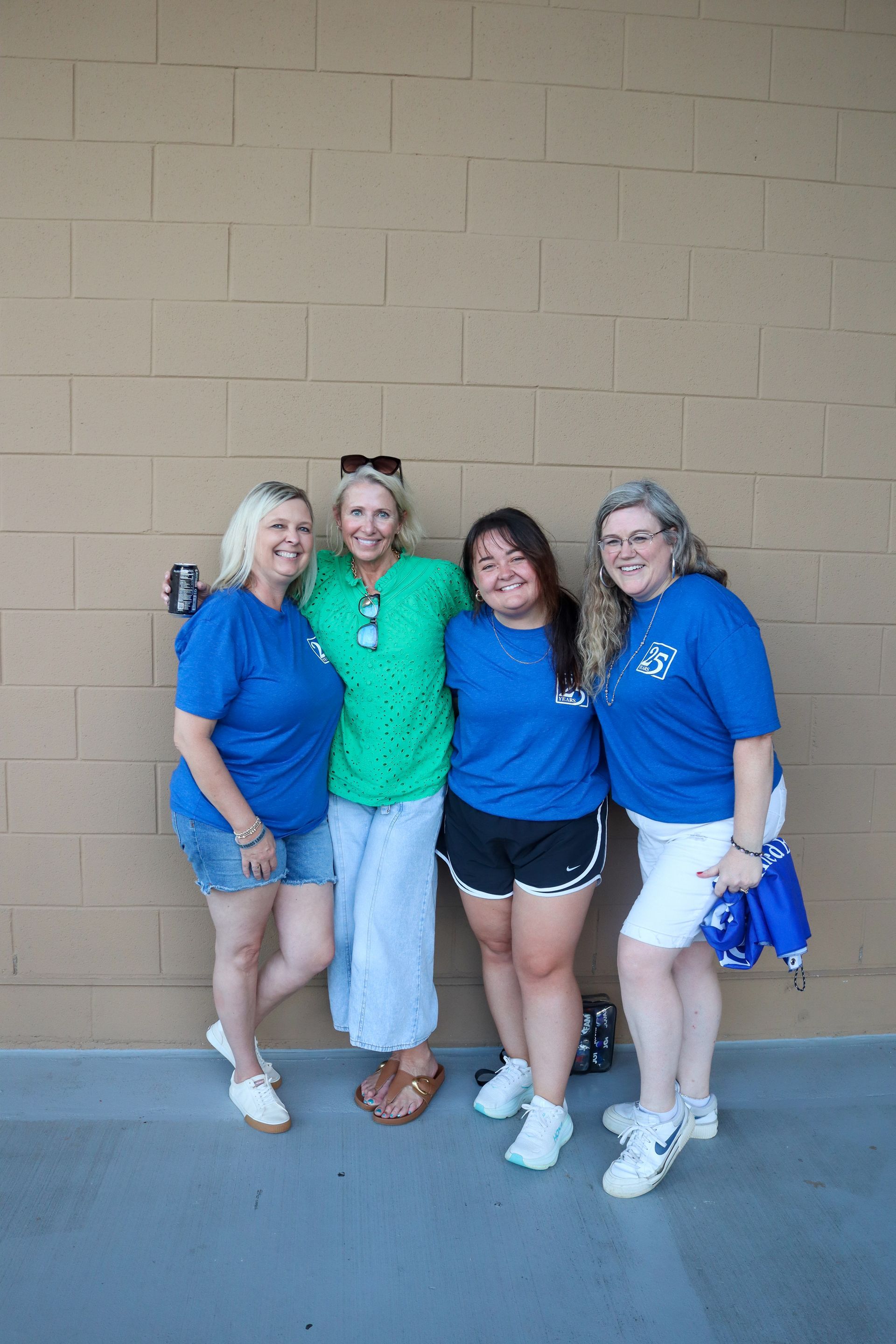 Four coworkers and friends standing together outdoors against a tan wall, wearing blue shirts and smiling.
