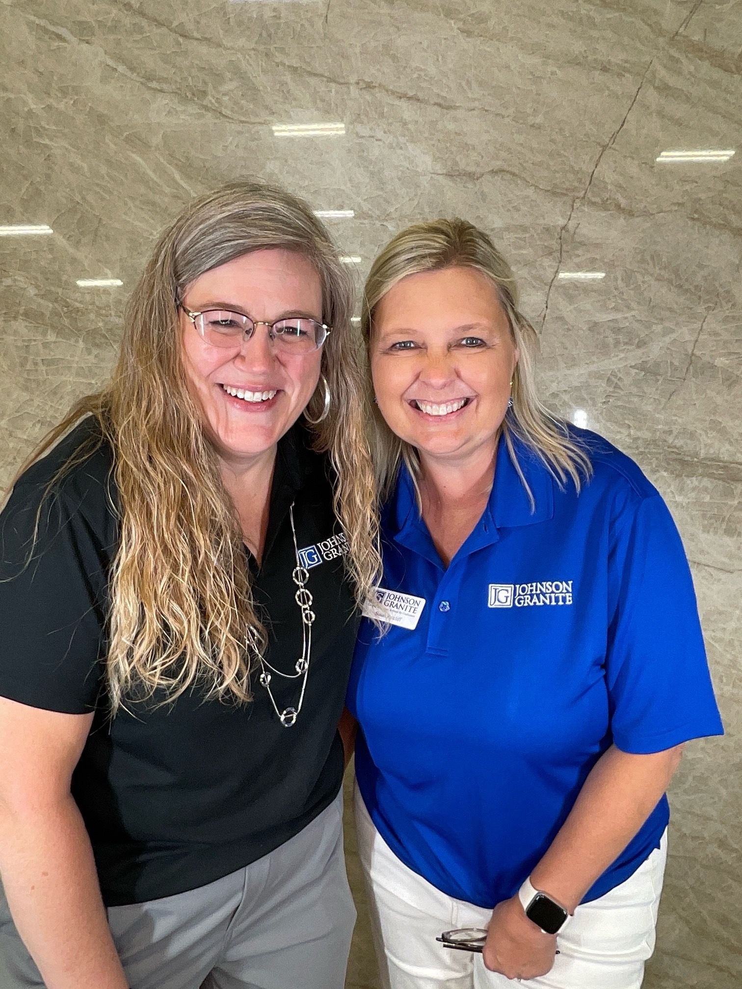 Two smiling women standing together indoors, one in a black shirt and one in a blue polo.