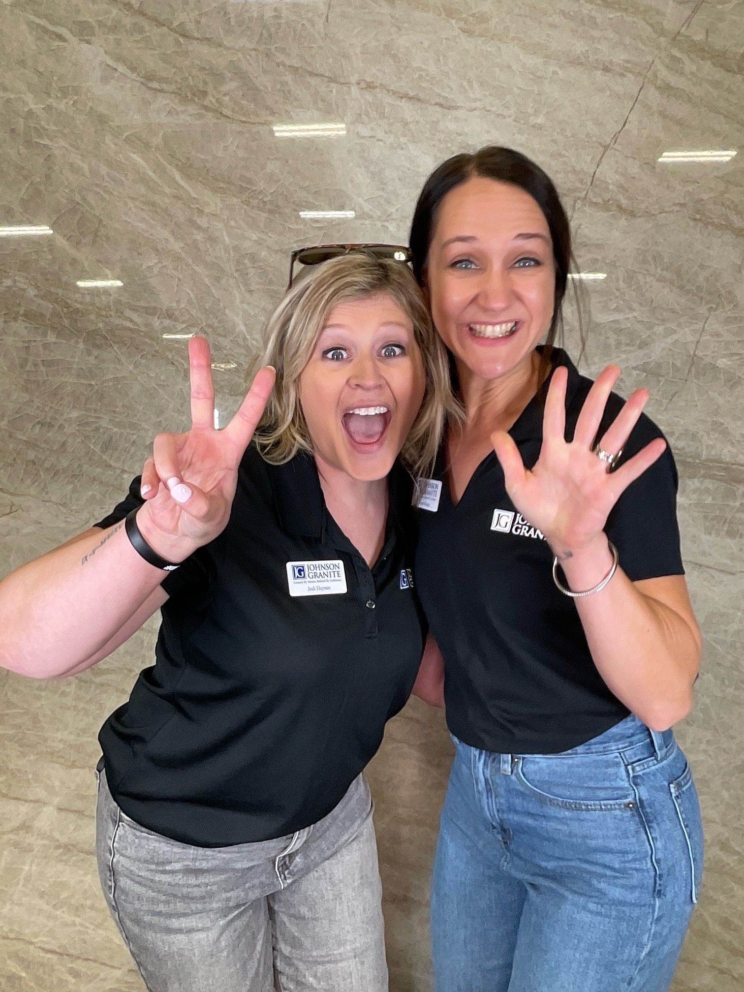 Two coworkers in black polos smiling and waving in a lobby with a stone wall background