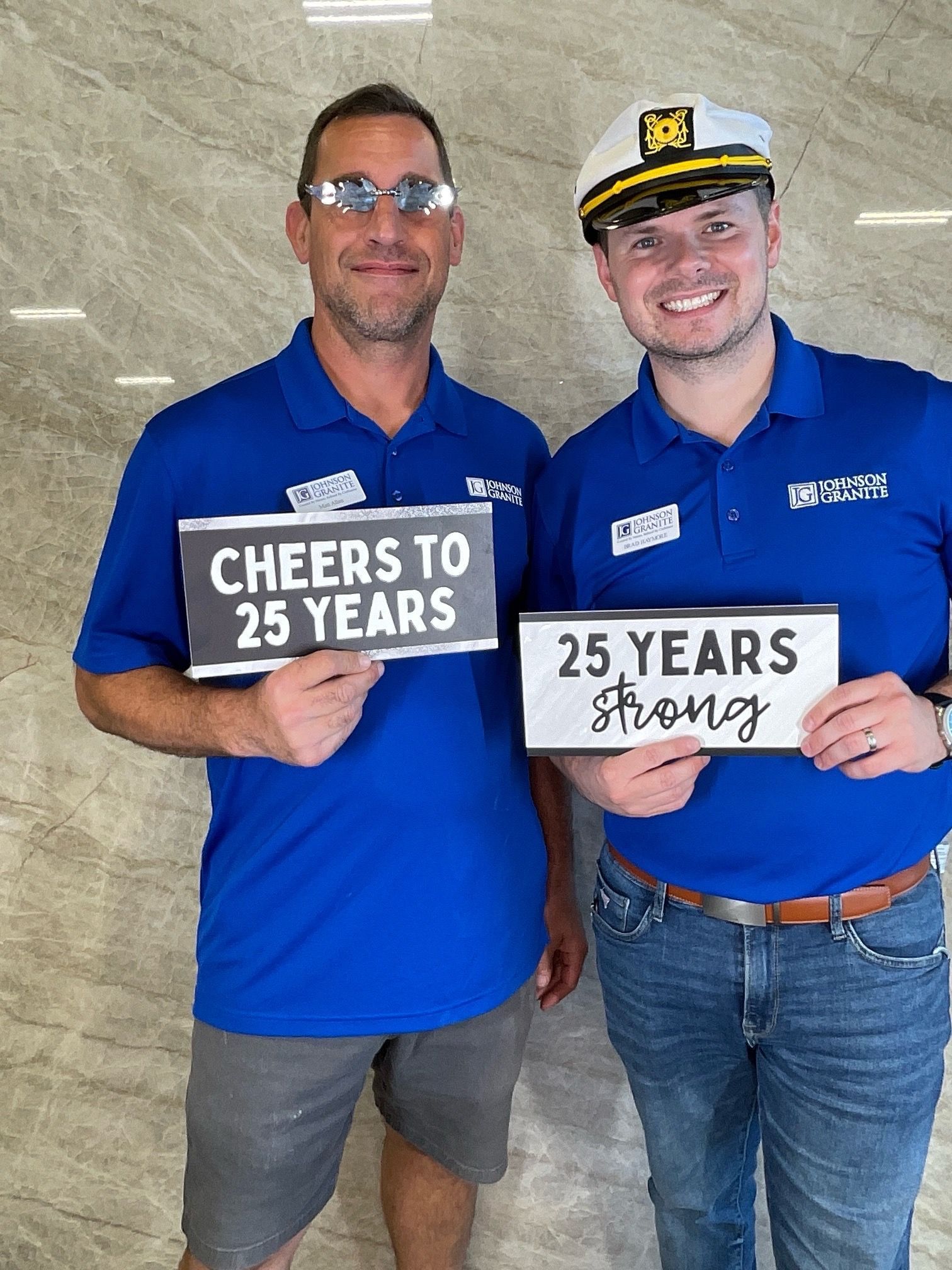 Two men in blue shirts holding signs that say “Cheers to 25 years” and “25 years strong”