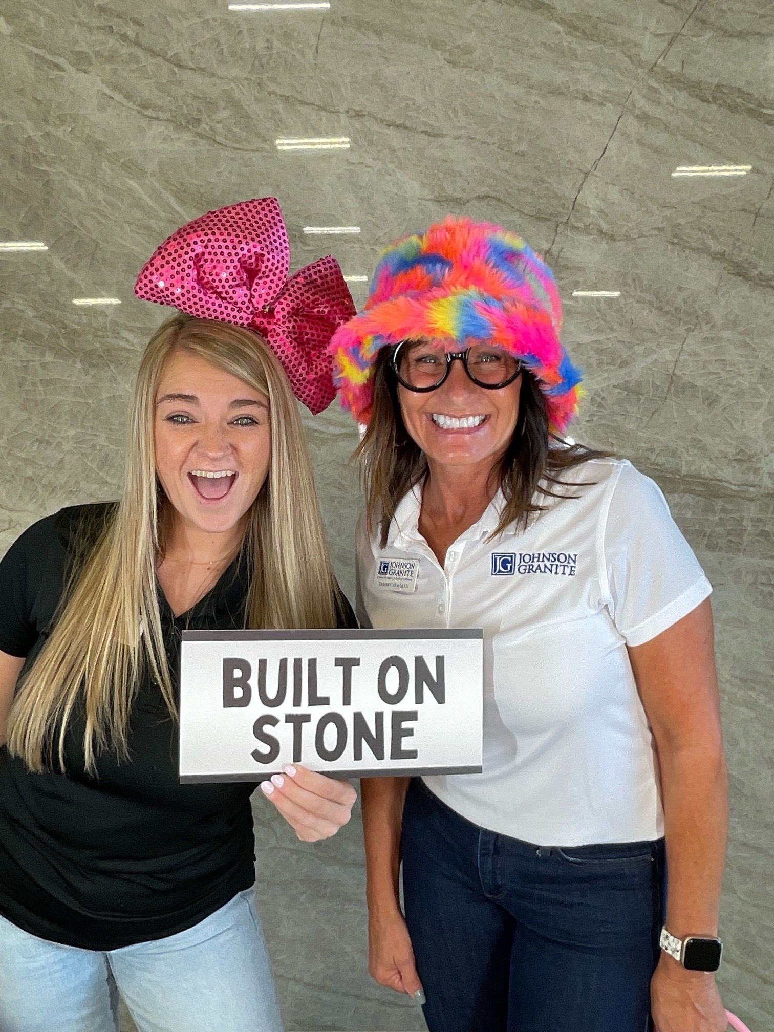 Two women wearing colorful hats hold a “BUILT ON STONE” sign and smile at the camera.