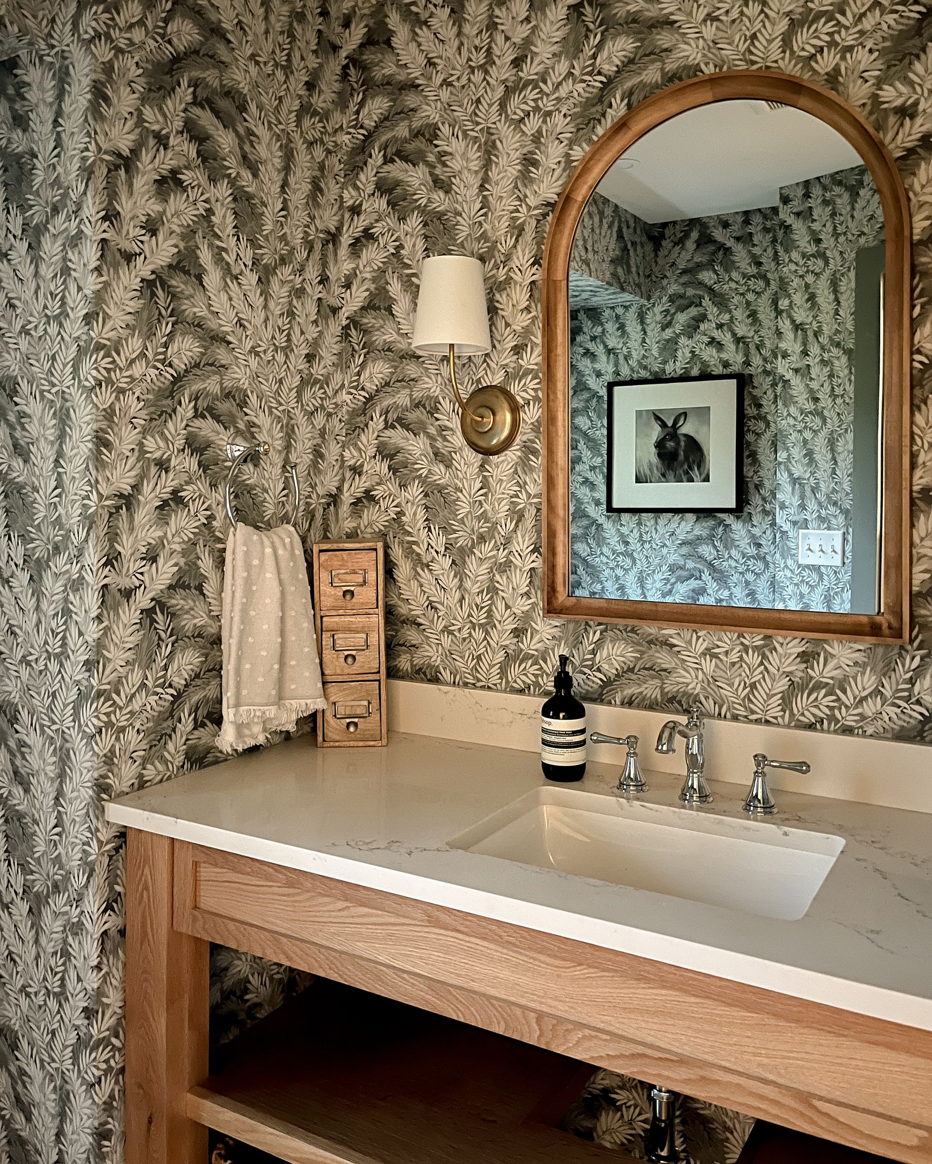 Bathroom vanity with beige countertop, wood-framed mirror, patterned wallpaper, and wall sconce.