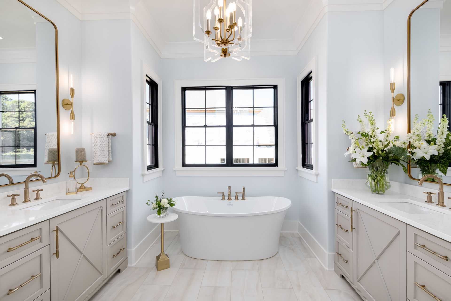 Bright, elegant bathroom with twin vanities, freestanding tub, large window, and white-and-gold decor
