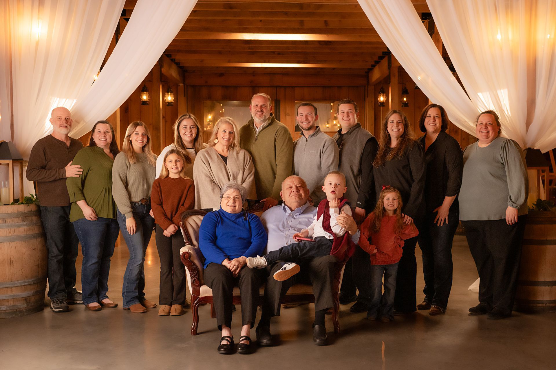 Group portrait of 16 people posed indoors in a rustic hall with draped white curtains and wooden walls