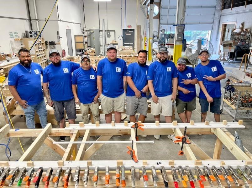 Group of workers in blue shirts posing in a workshop beside a large wooden frame and machinery
