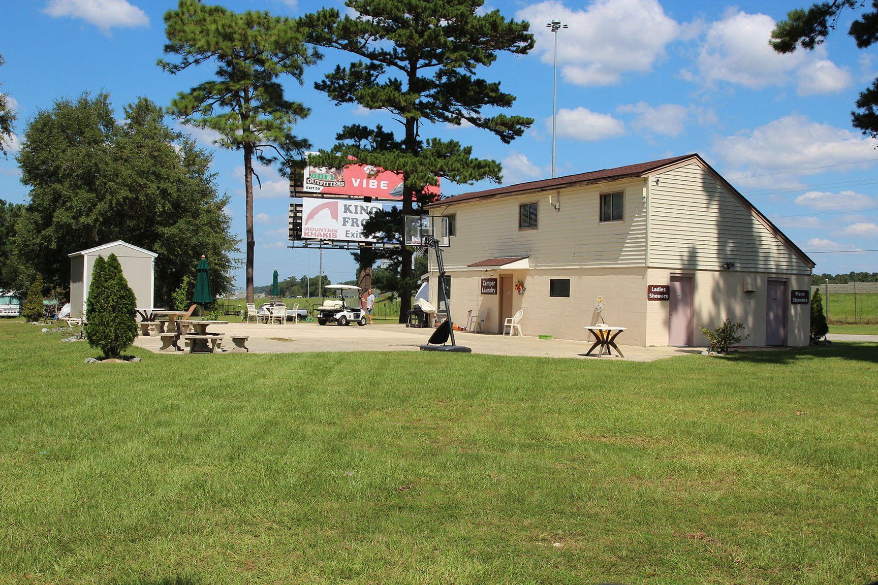Building with a grassy yard. Trees, a billboard, and outdoor seating are visible. Blue sky.