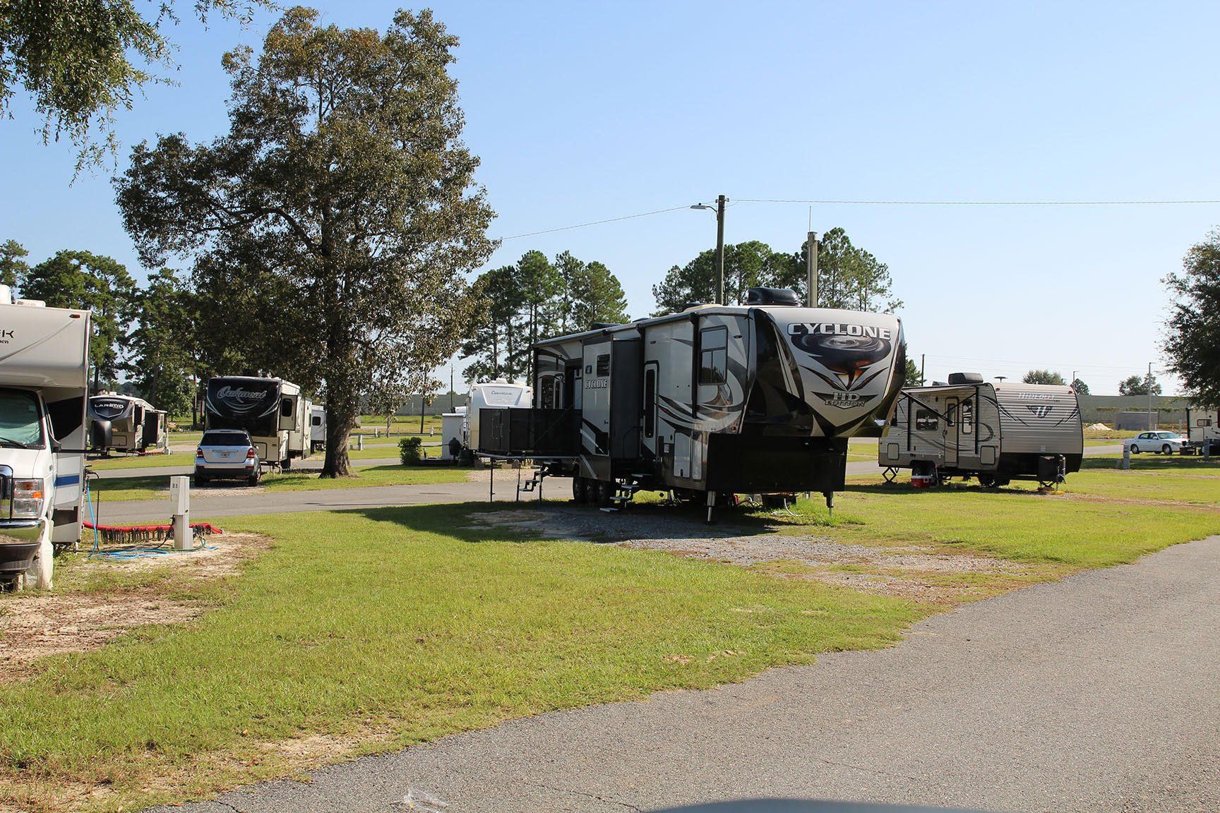Campground with RVs parked on grass, gravel road, trees, and blue sky.