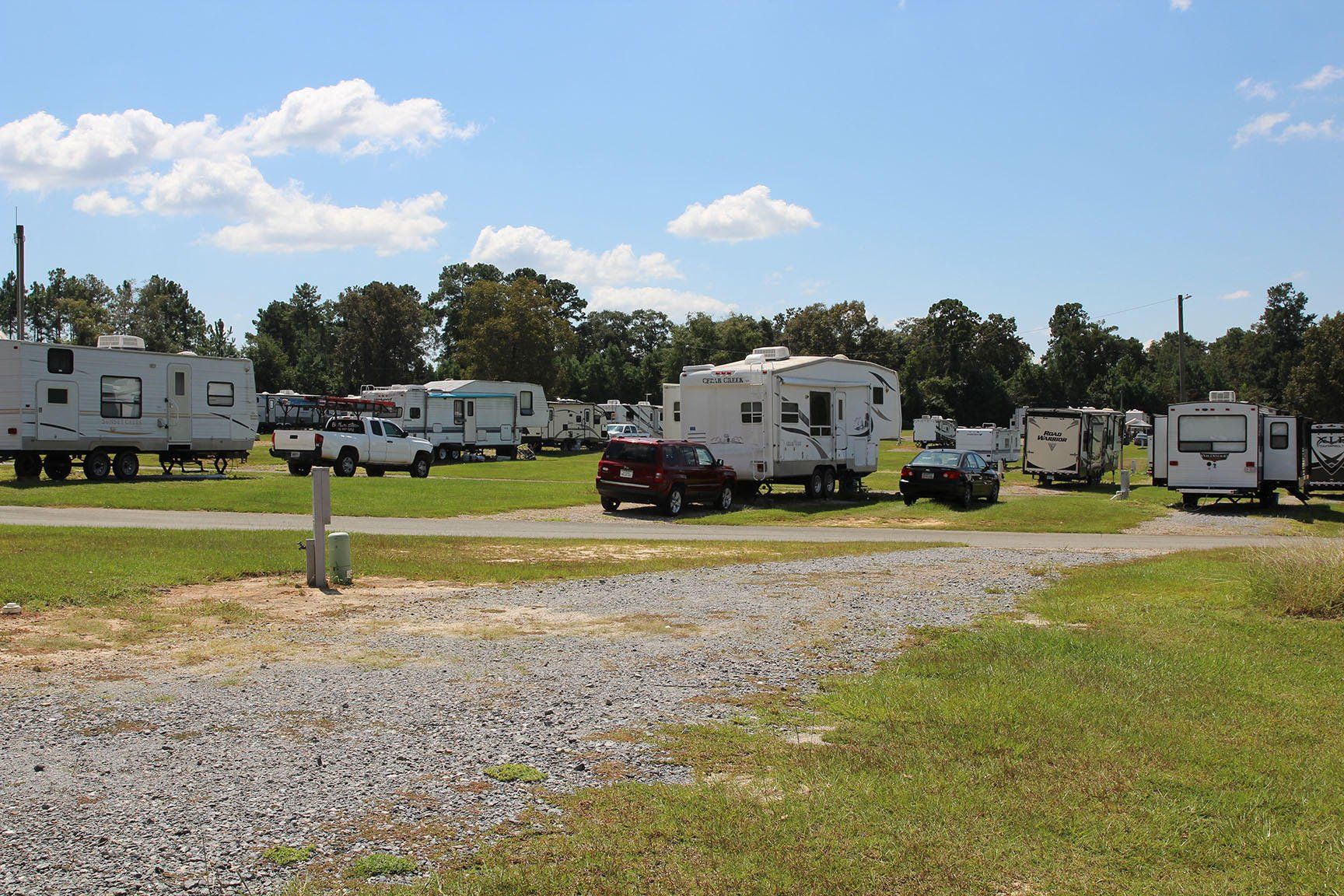 RV campground with gravel road and grassy areas; sunny day with white clouds.