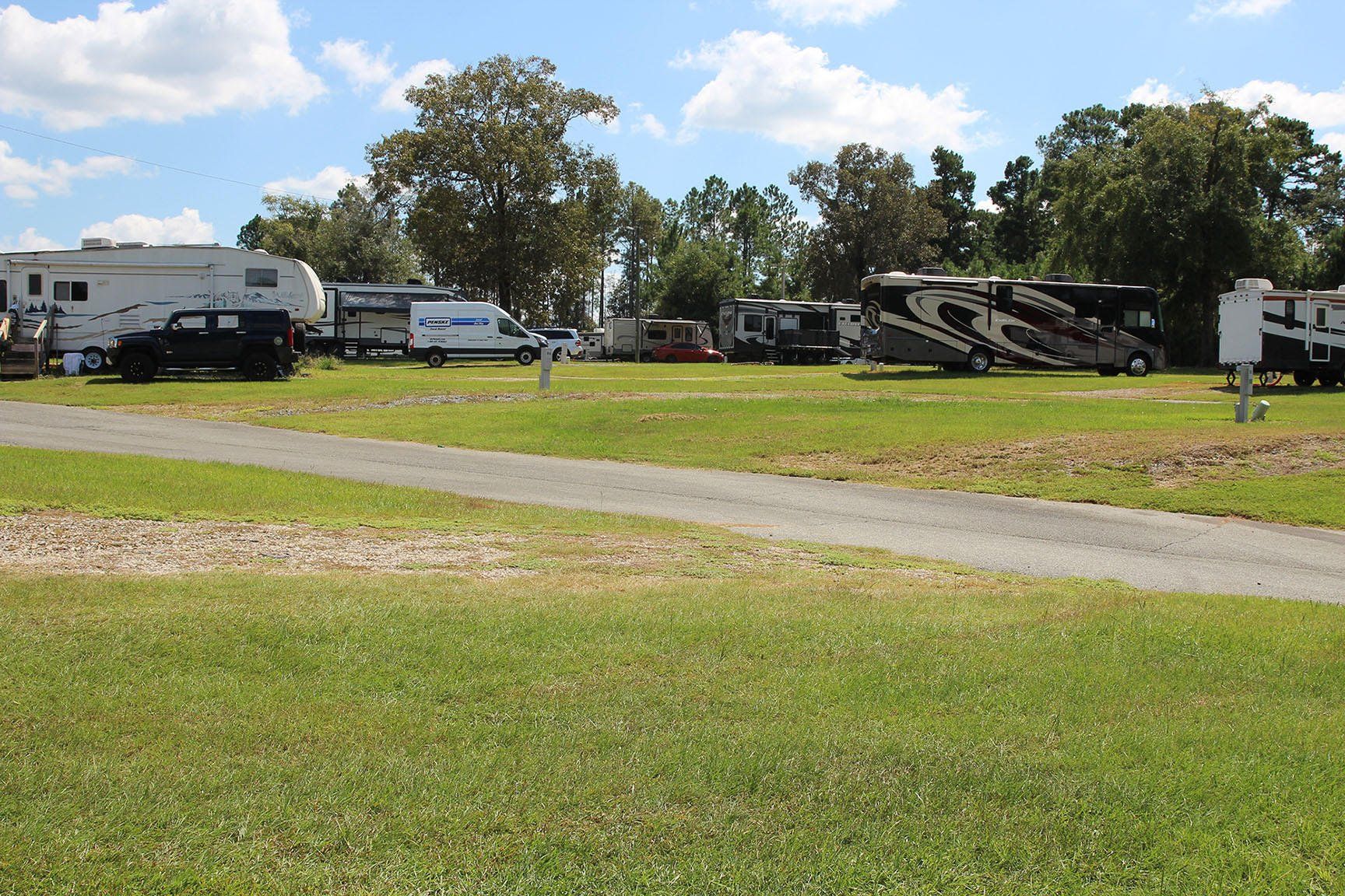 RV campground with RVs parked on grass, gravel path, and trees in background under a cloudy sky.