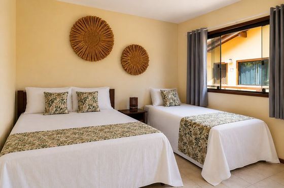 Duas camas impecavelmente arrumadas em um quarto bege claro com decoração de parede em tecido e vista para a janela.
