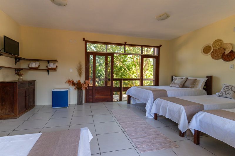 Quarto de hóspedes luminoso com duas camas de solteiro, piso de azulejo, móveis de madeira e grandes janelas com vista para a vegetação.
